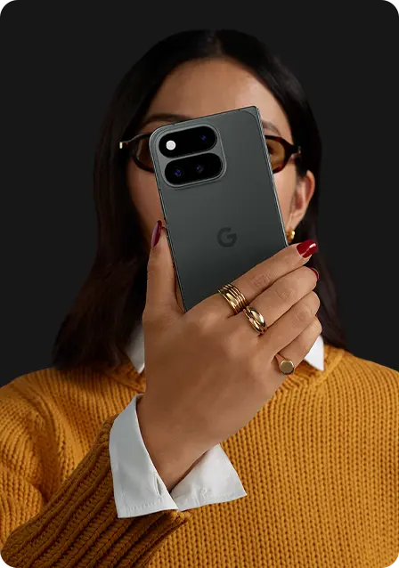 Person holding a smartphone, wearing glasses and a sweater, against a dark background.