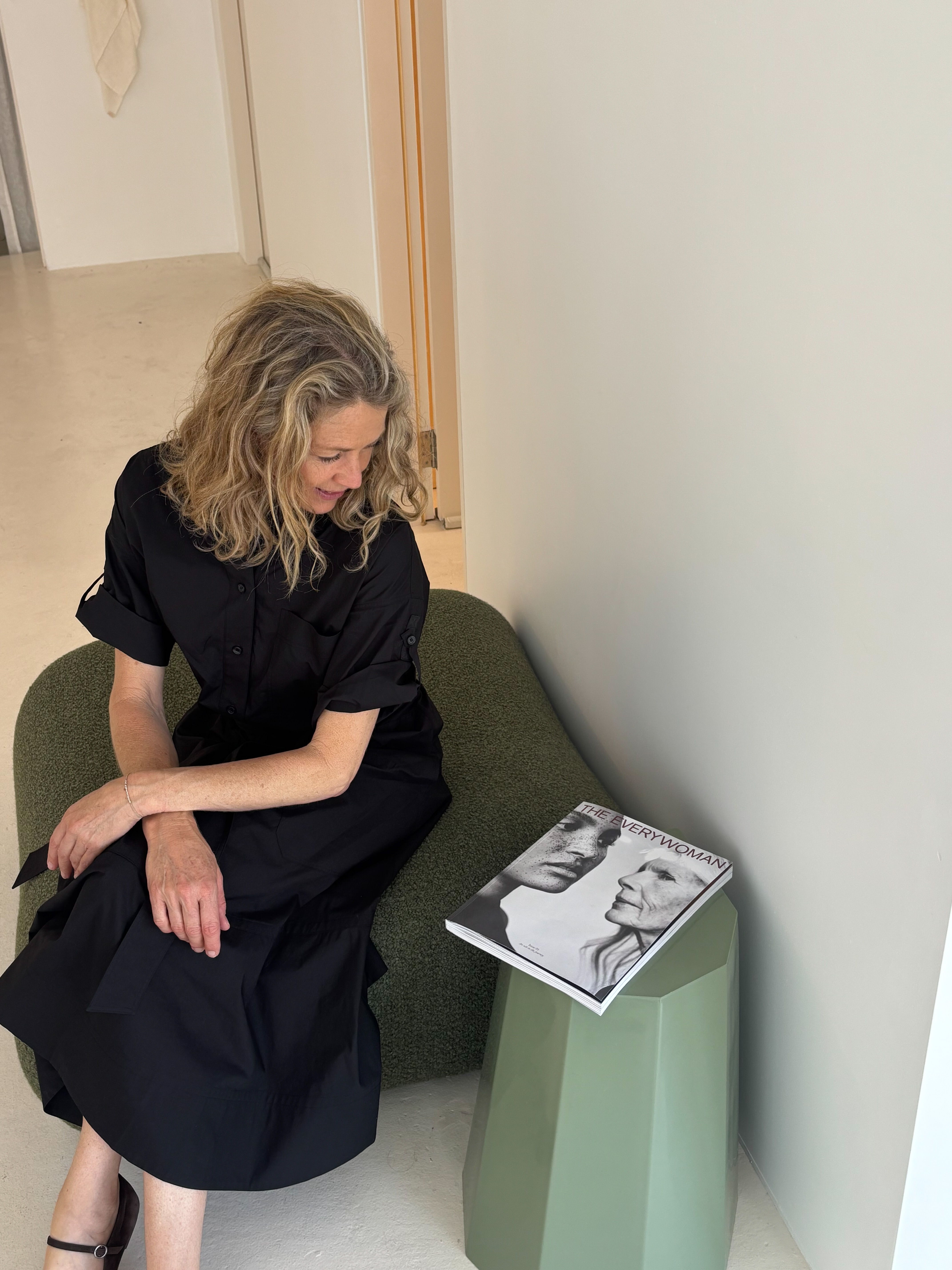 A woman in a black dress sits beside a green table, reading a magazine.