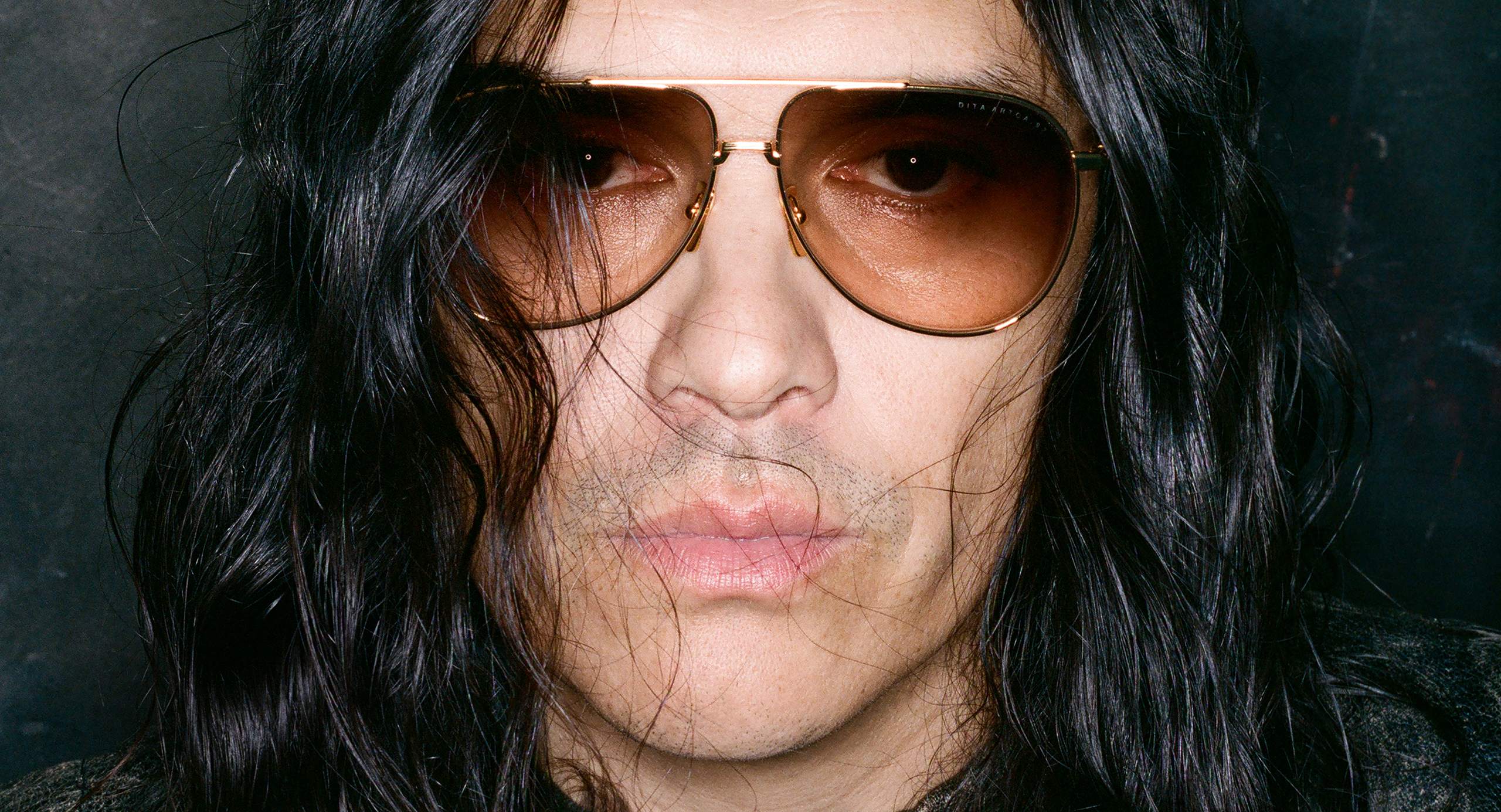 Guillermo Andrade, with long black hair wearing sunglasses against a dark background.