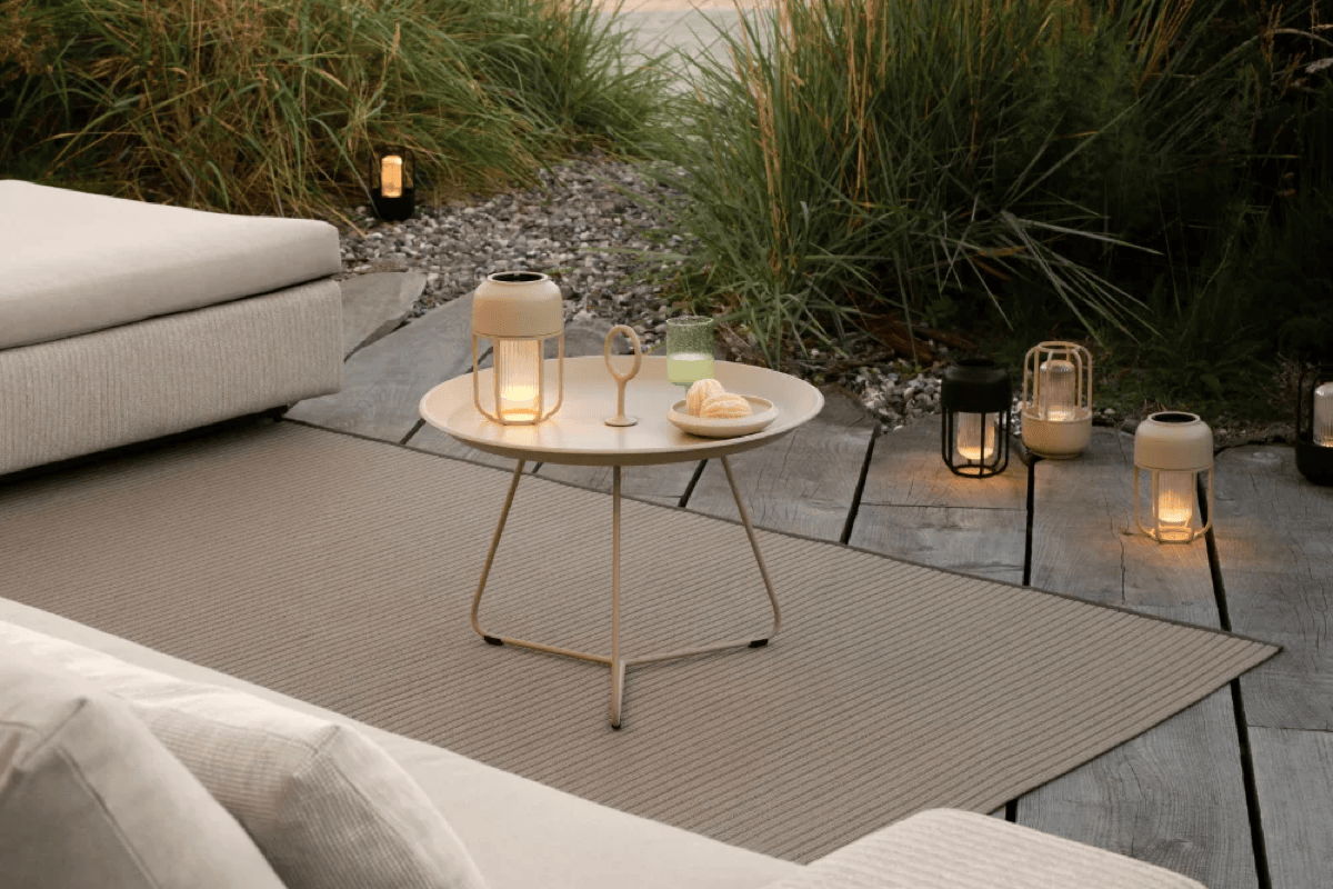 Outdoor side table with lanterns and a drink on a neutral rug at dusk, with small path lights glowing in the landscape.