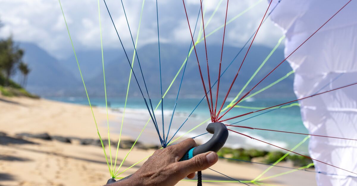 Person holding a colorful parachute on a beach with mountains in the background.