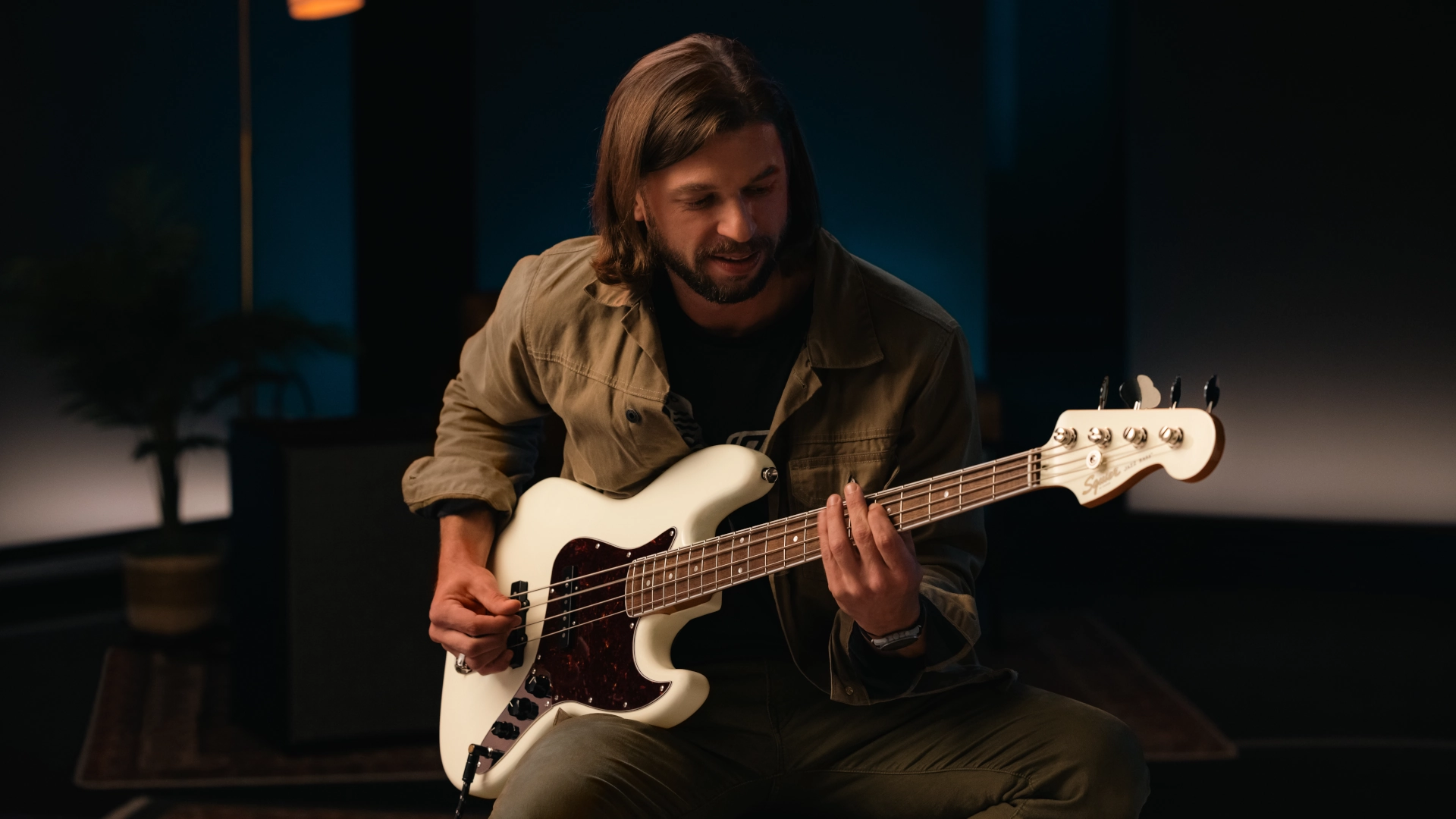 Man playing a white Squier Jazz Bass guitar in a dimly lit room.
