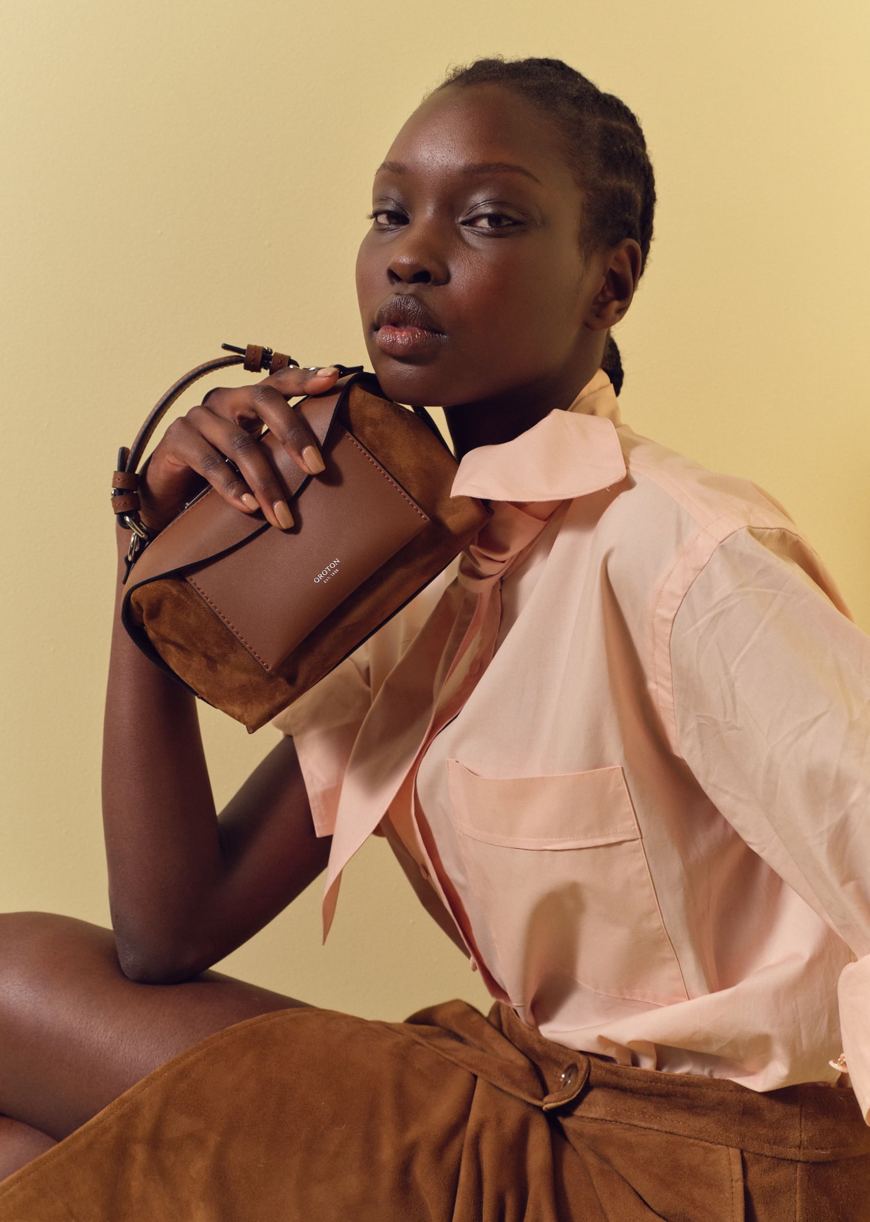 Model in a pink shirt holding a brown handbag against a yellow background.