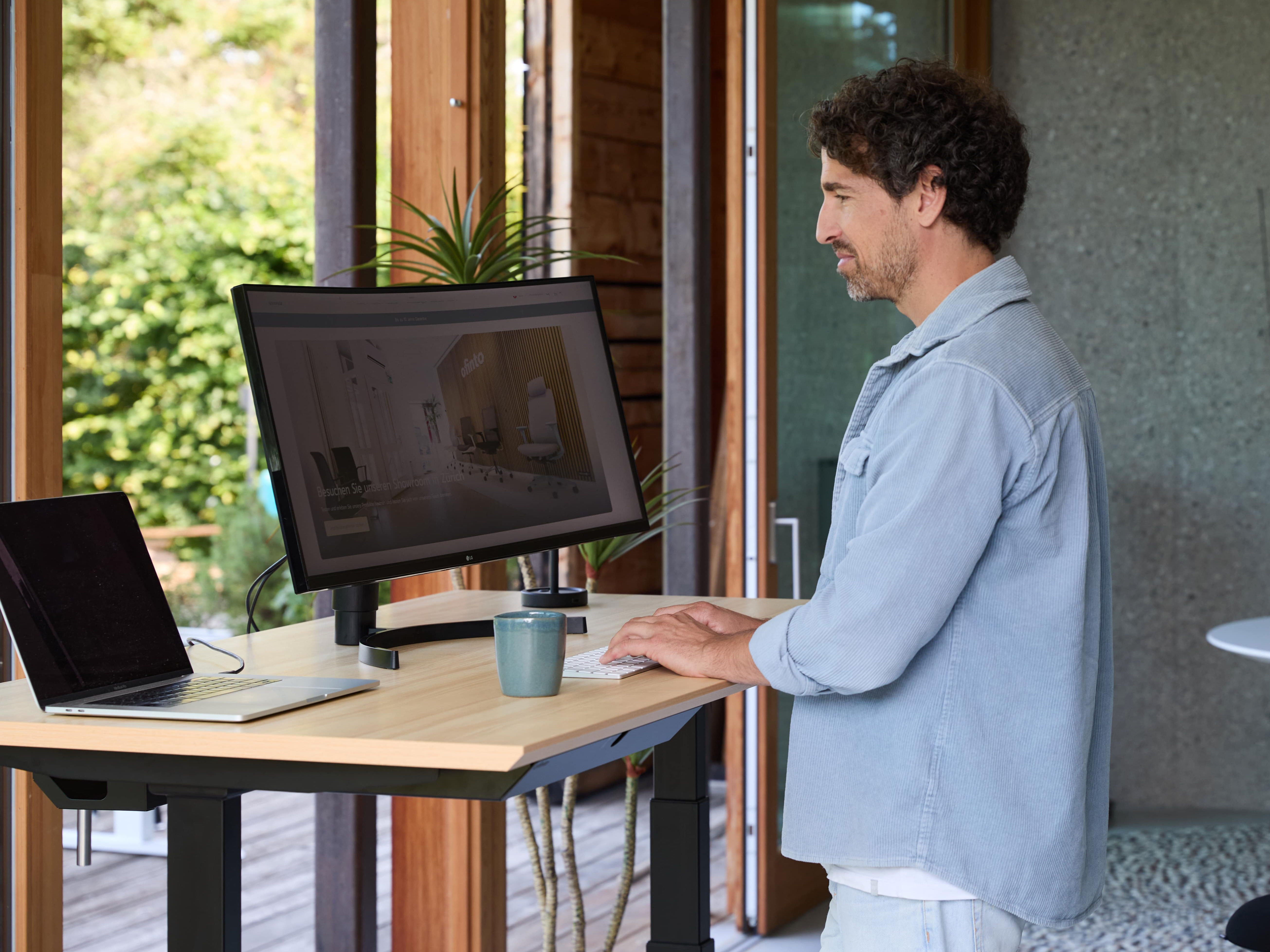 Man works concentrated at the height-adjustable standing desk with monitor and laptop, modern and natural environment with lots of daylight.