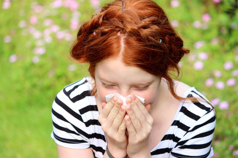 Junges Mädchen, das an einem sonnigen Nachmittag in einem Garten sitzt. Sie niest und schnäuzt sich die Nase, da sie an Heuschnupfen leidet.