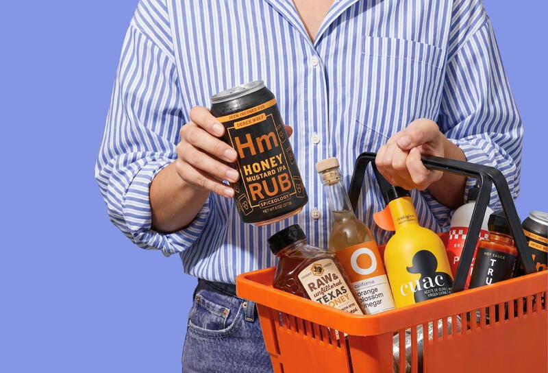Person holding a Honey Mustard IPA Rub can and an orange shopping basket filled with artisanal sauces and honey.