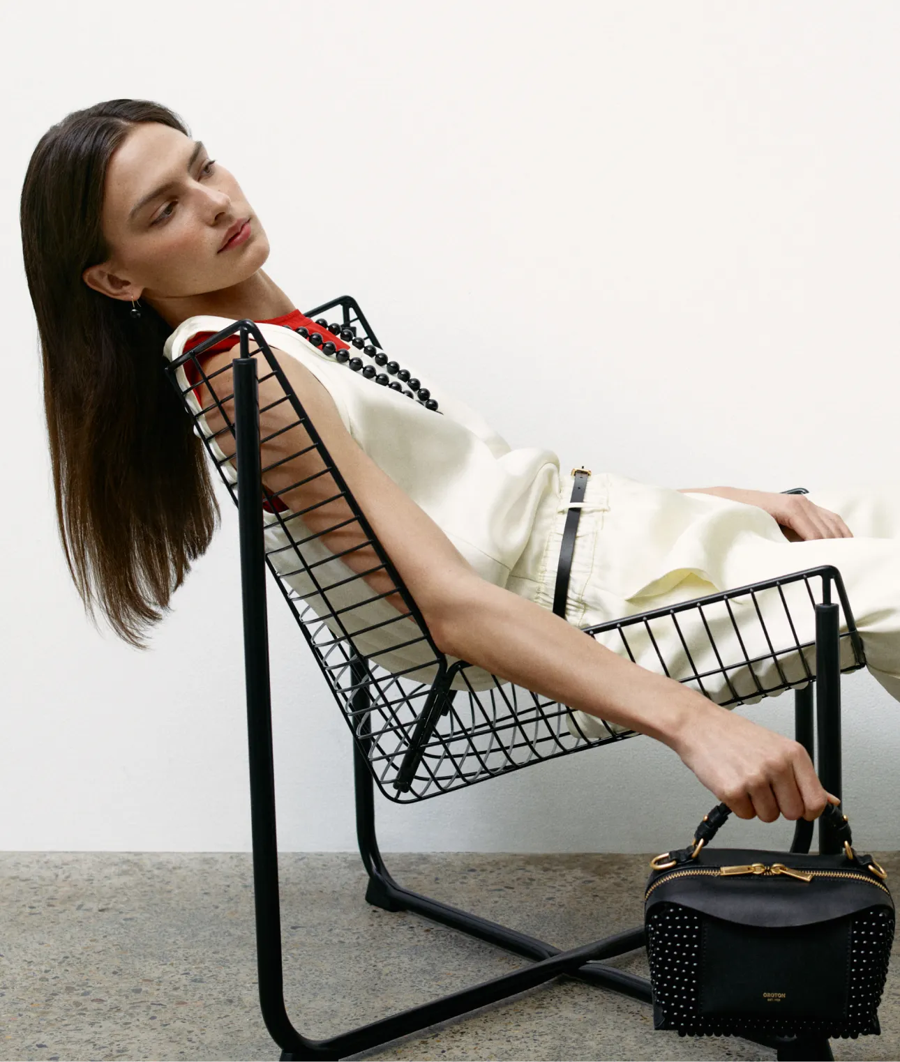 Model reclining in a modern wire chair, holding a stylish handbag.