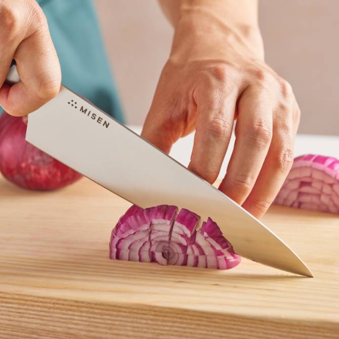 Hands use a blue Misen Chef’s Knife to dice red onions on a light wood cutting board.
