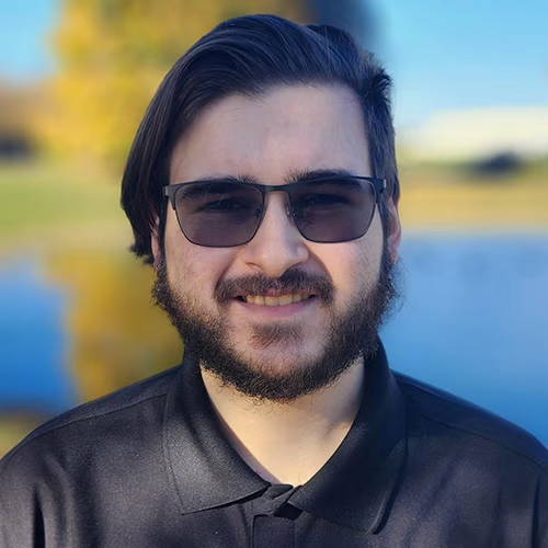 Person with sunglasses smiling outdoors near water and trees.