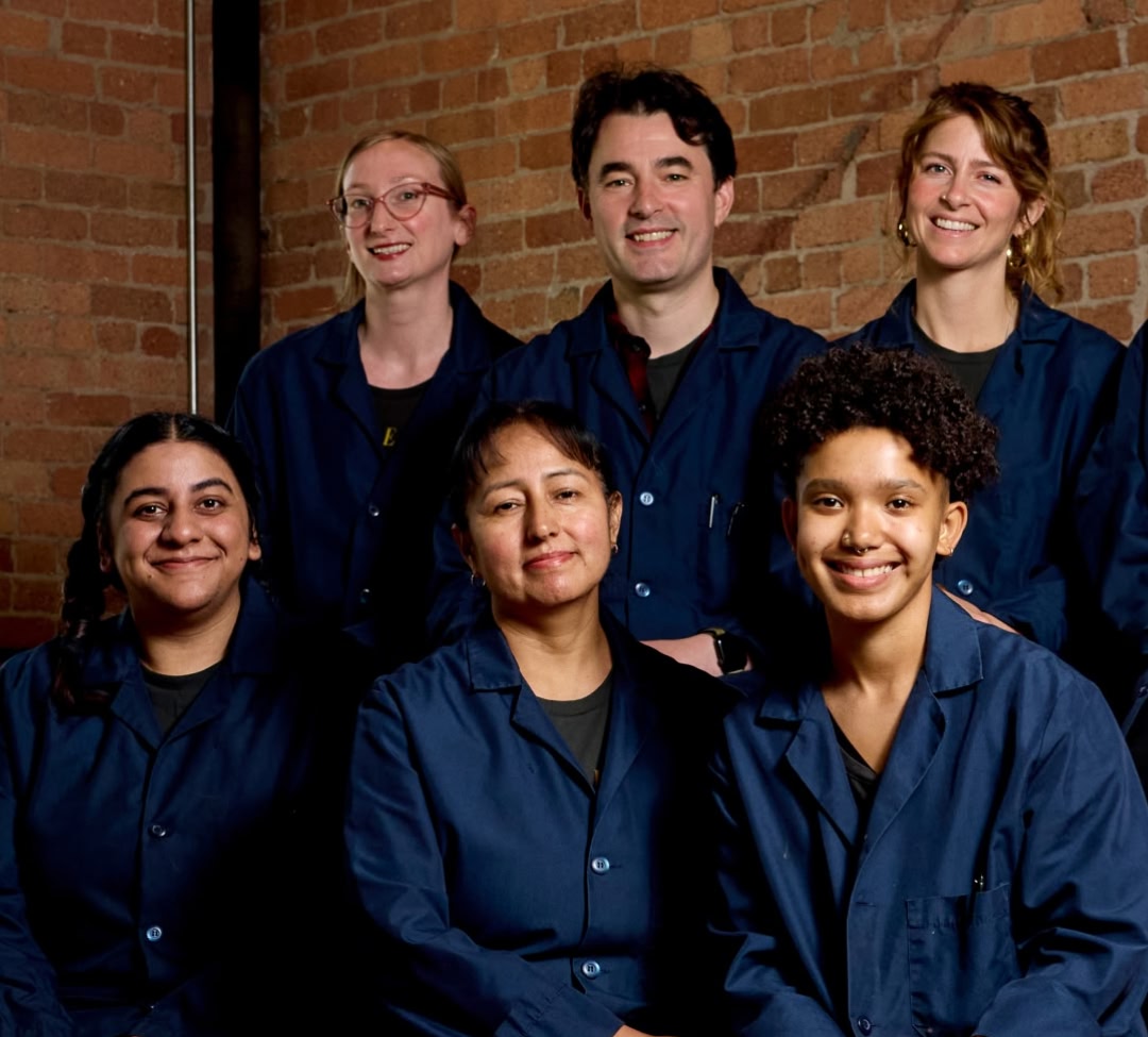 A group of six people wearing blue lab coats, standing against a brick wall.