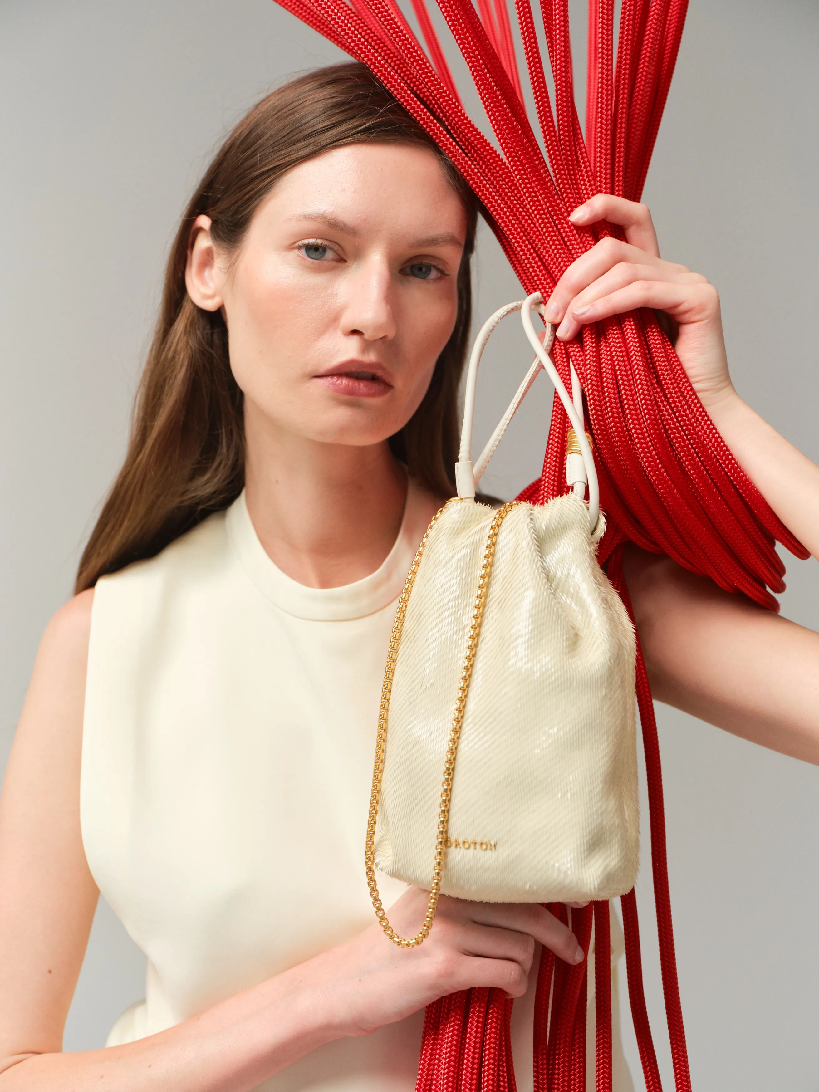 Model holding a cream handbag with red decorative elements in the background.