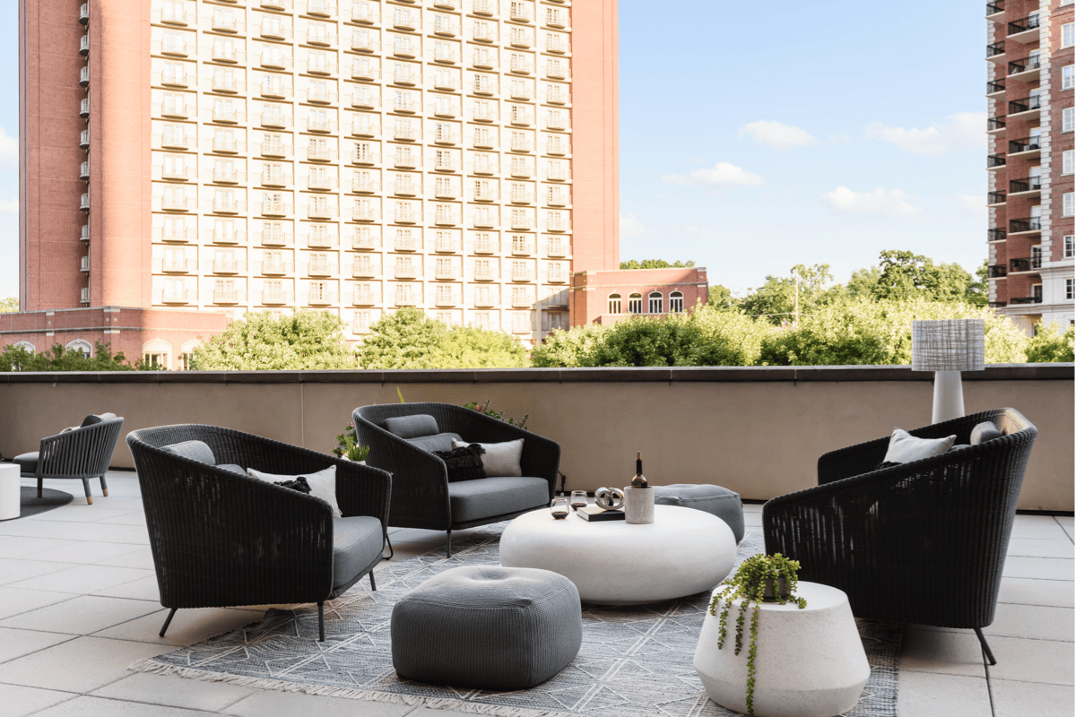 Rooftop outdoor lounge with woven club chairs, upholstered ottomans, and round pebble-style side tables on a patterned outdoor rug.