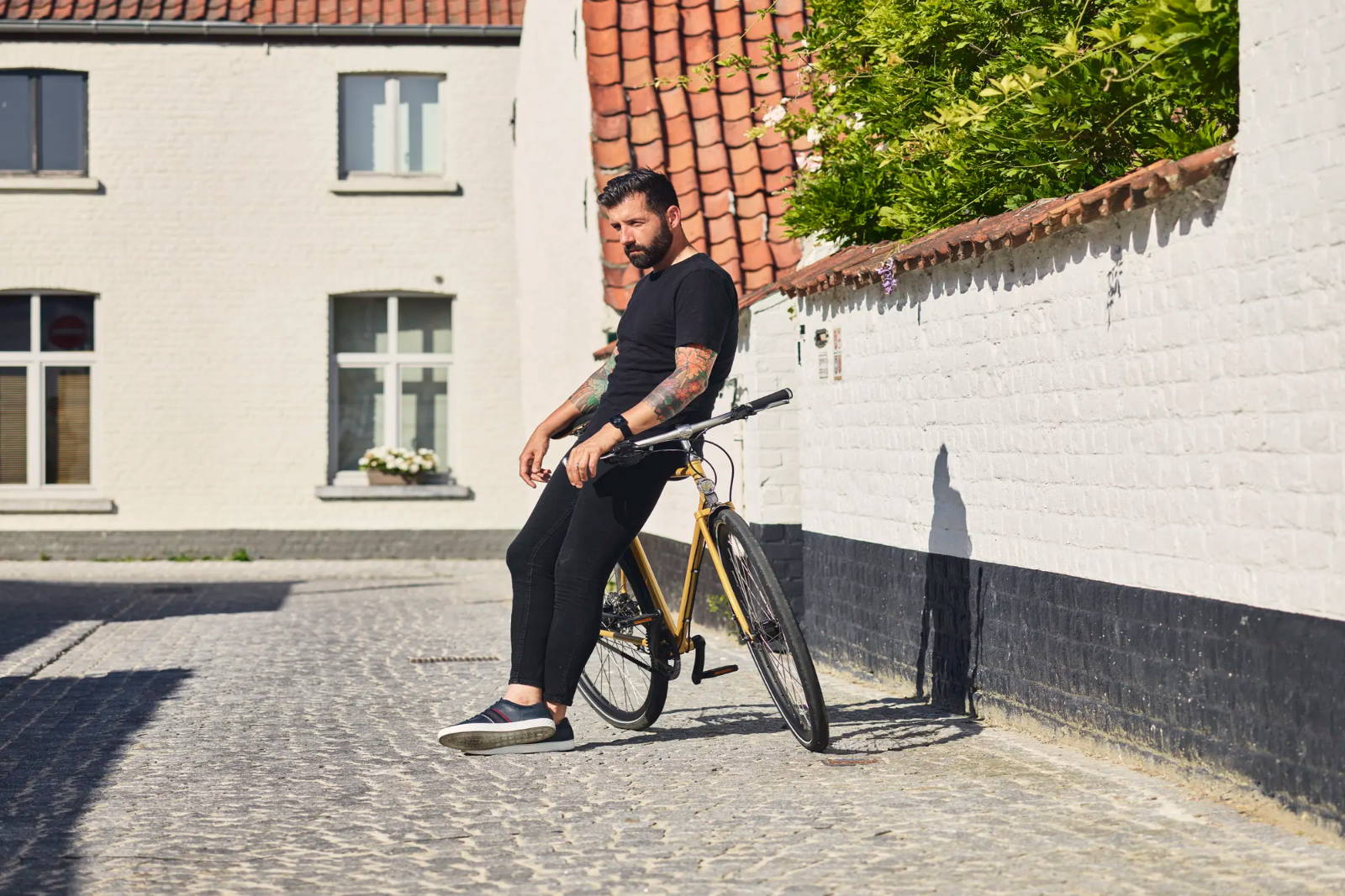 A man wearing all black leans against a mustard gold Achielle Sam bike in a cobblestone courtyard.