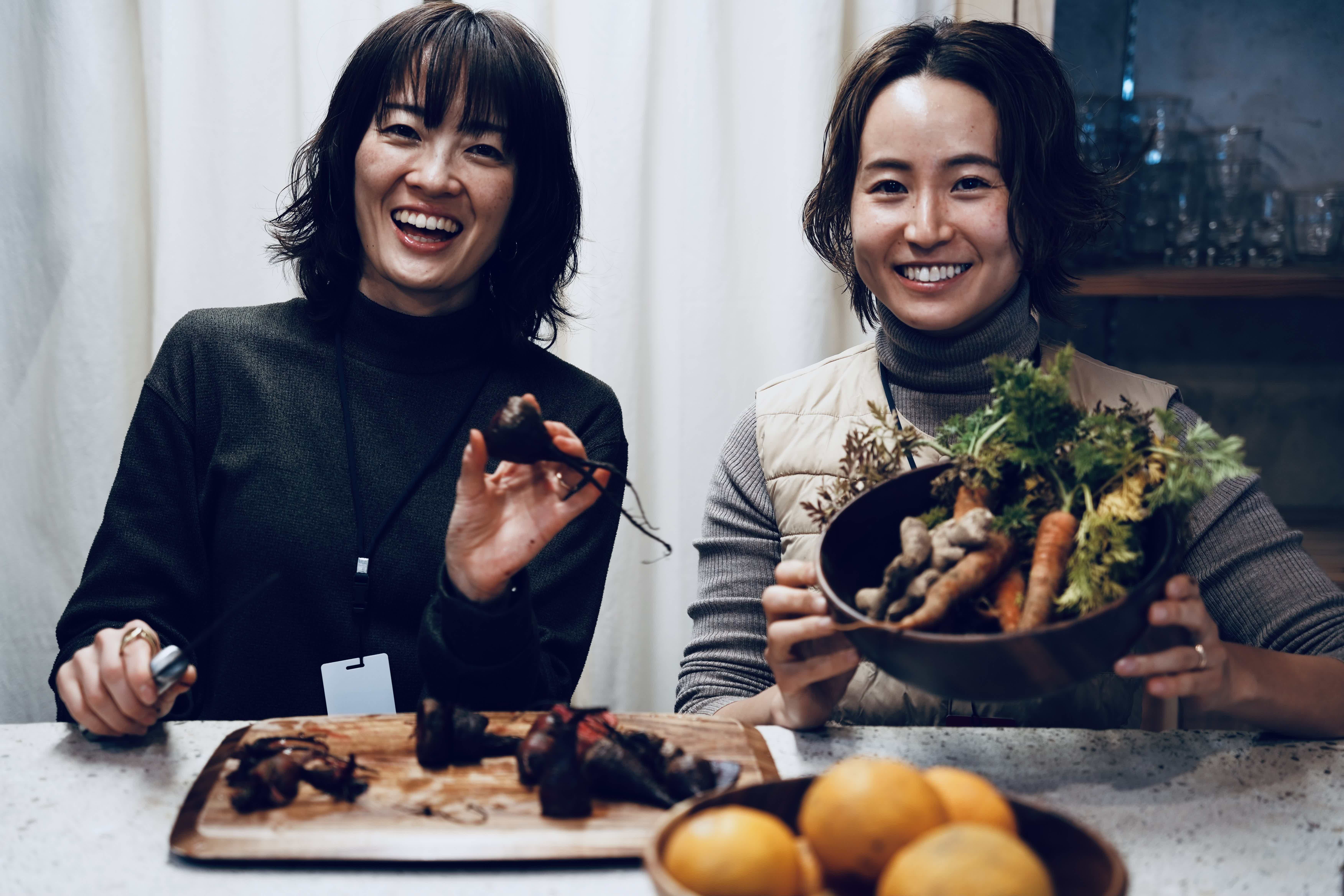 Two women smiling, holding vegetables, in a kitchen setting.