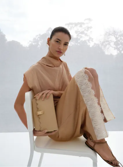A woman in a stylish beige outfit sitting on a chair, holding a clutch.