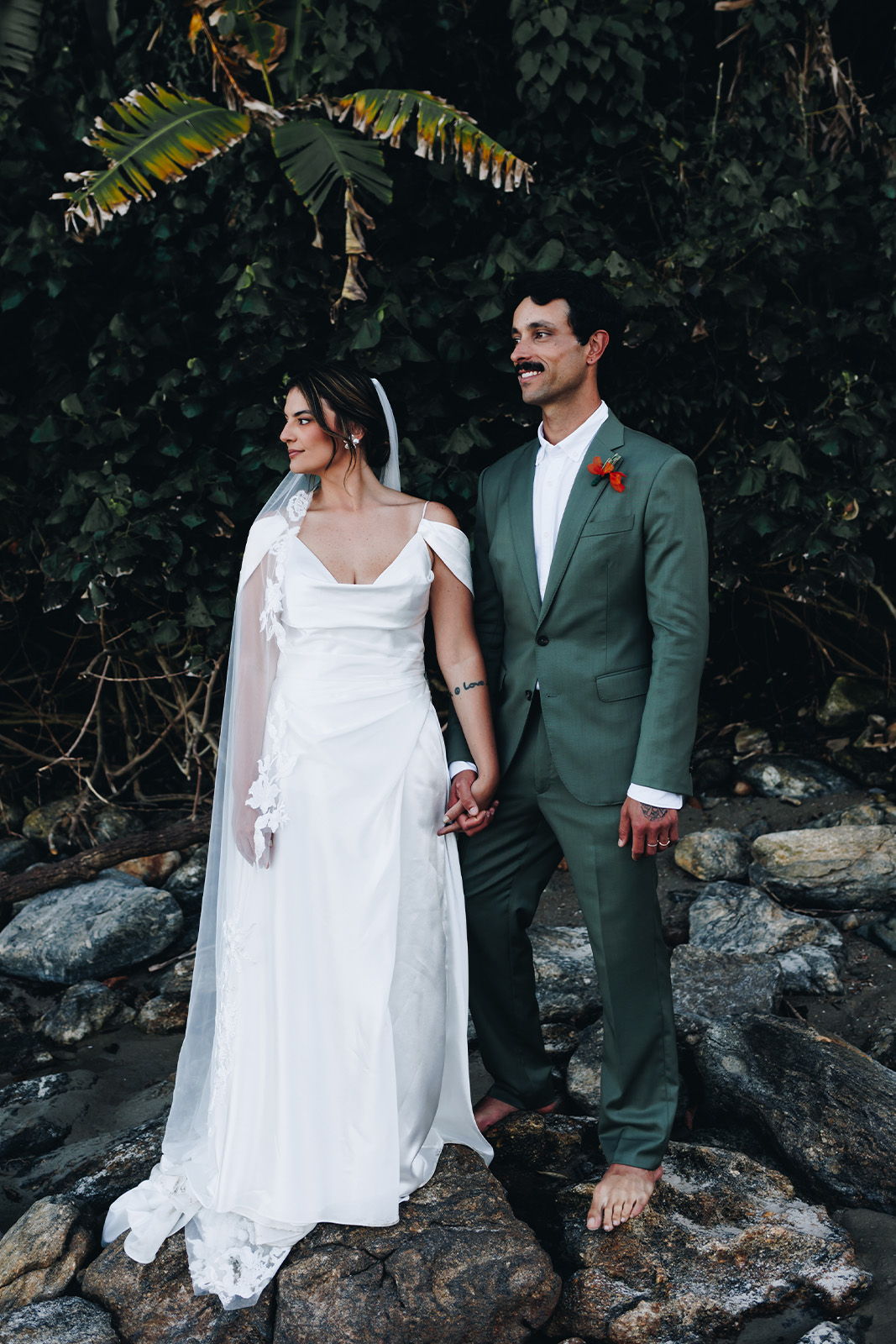 Couple in wedding attire standing on rocks near greenery.
