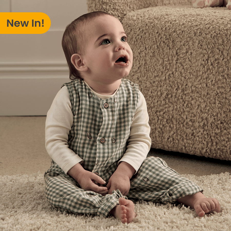 A baby sitting on a carpet, wearing a checkered outfit, looking up.