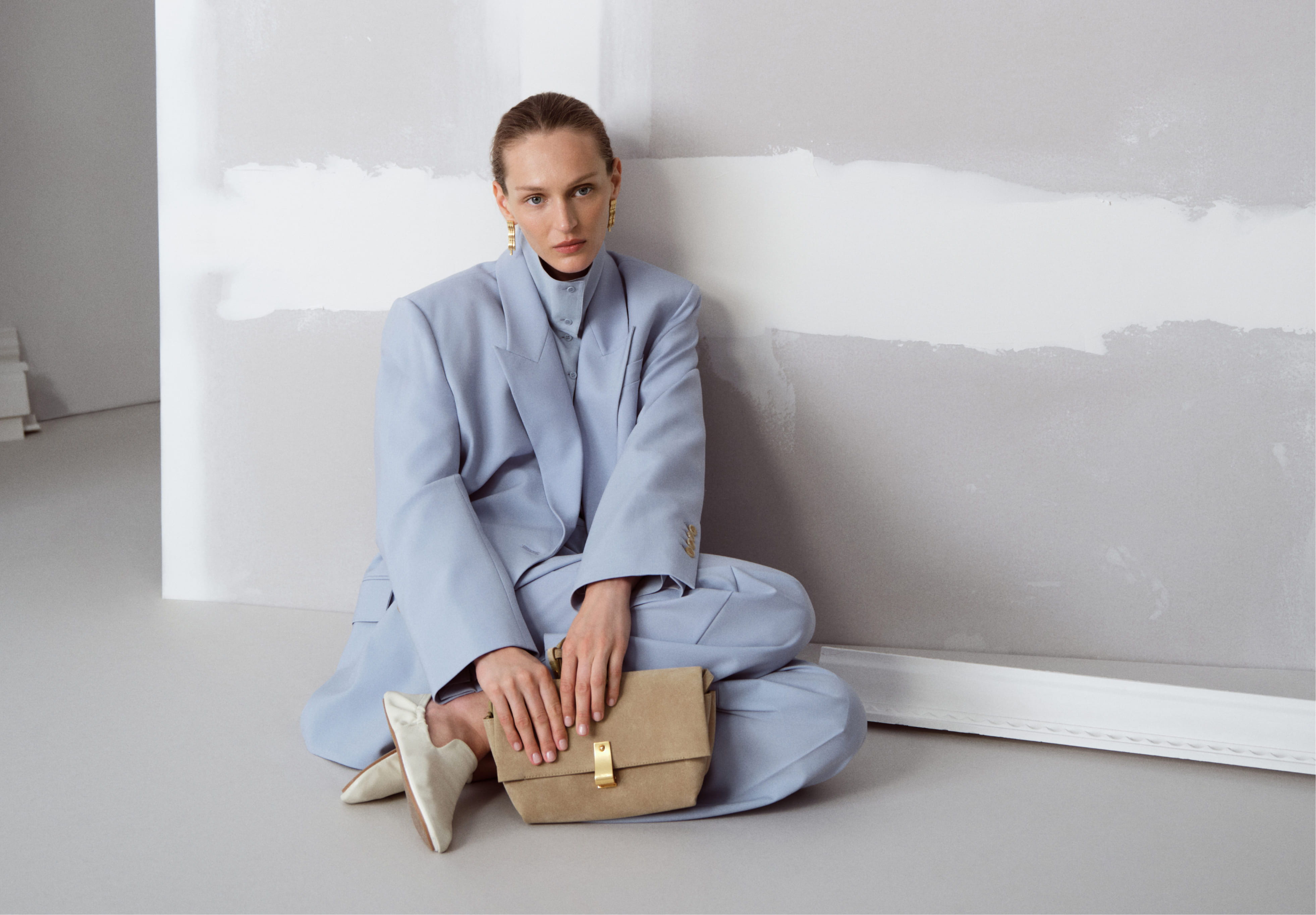Person sitting against a wall, wearing a light blue suit, holding a bag.