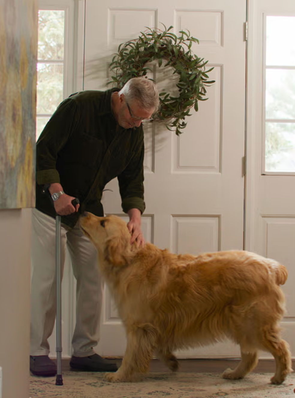 Older man using a single Y-Cane while bending down to pet a golden retriever near a front door