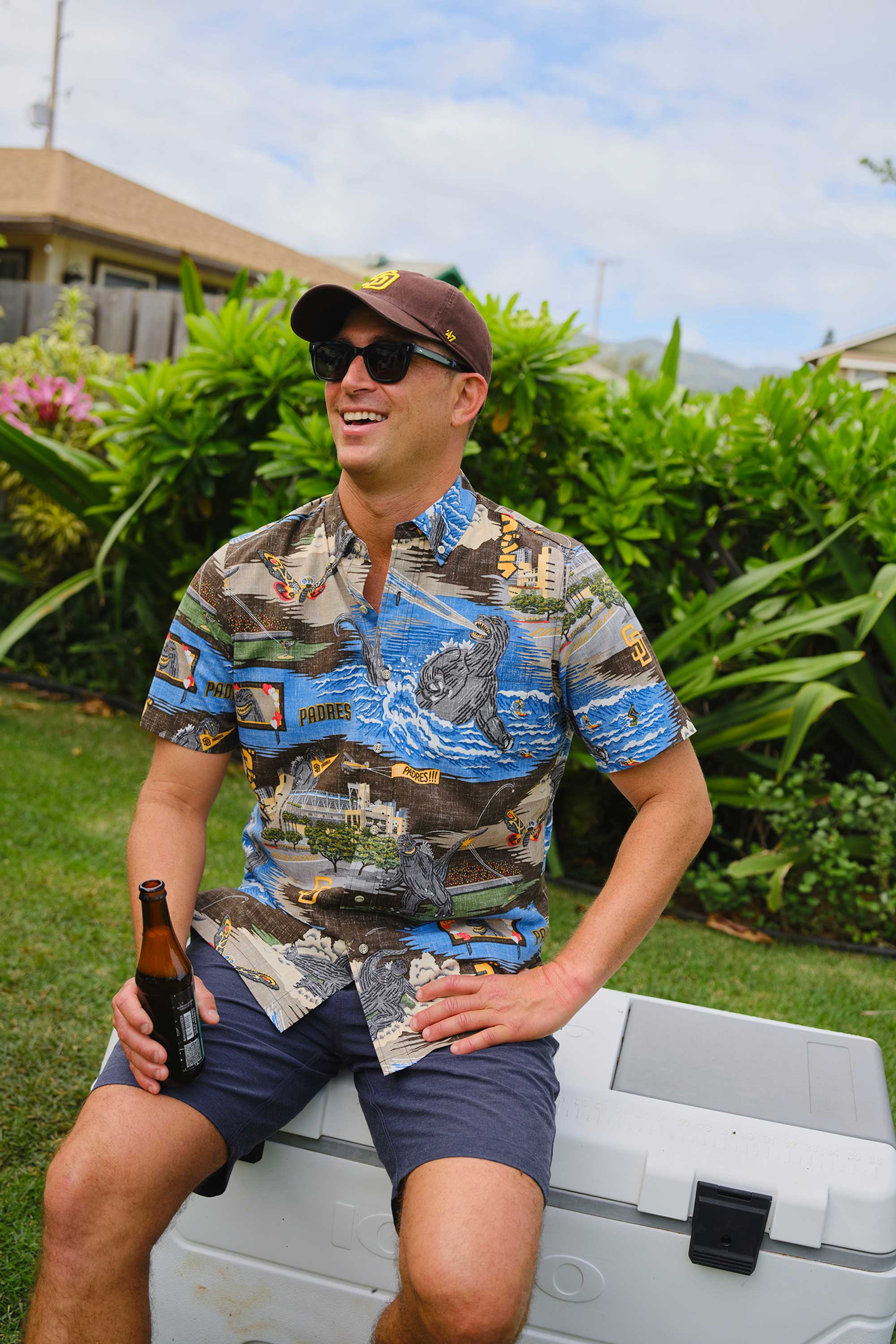 Man in a colorful shirt sitting on a cooler, holding a beer.