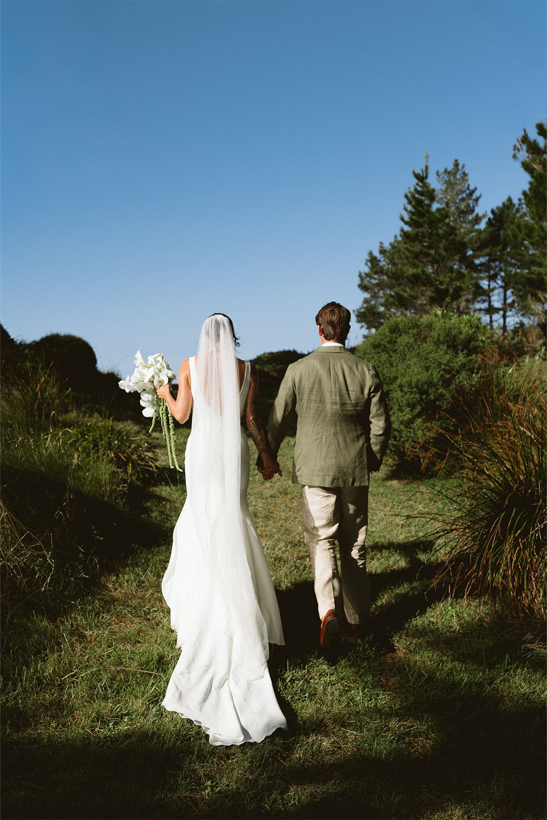 Couple walking hand-in-hand through a lush outdoor setting, bride in white dress.