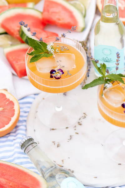 Peach mocktail in coupe glass, topped with pansies and herbs. It is placed on a tray with watermelon and grapefruit.