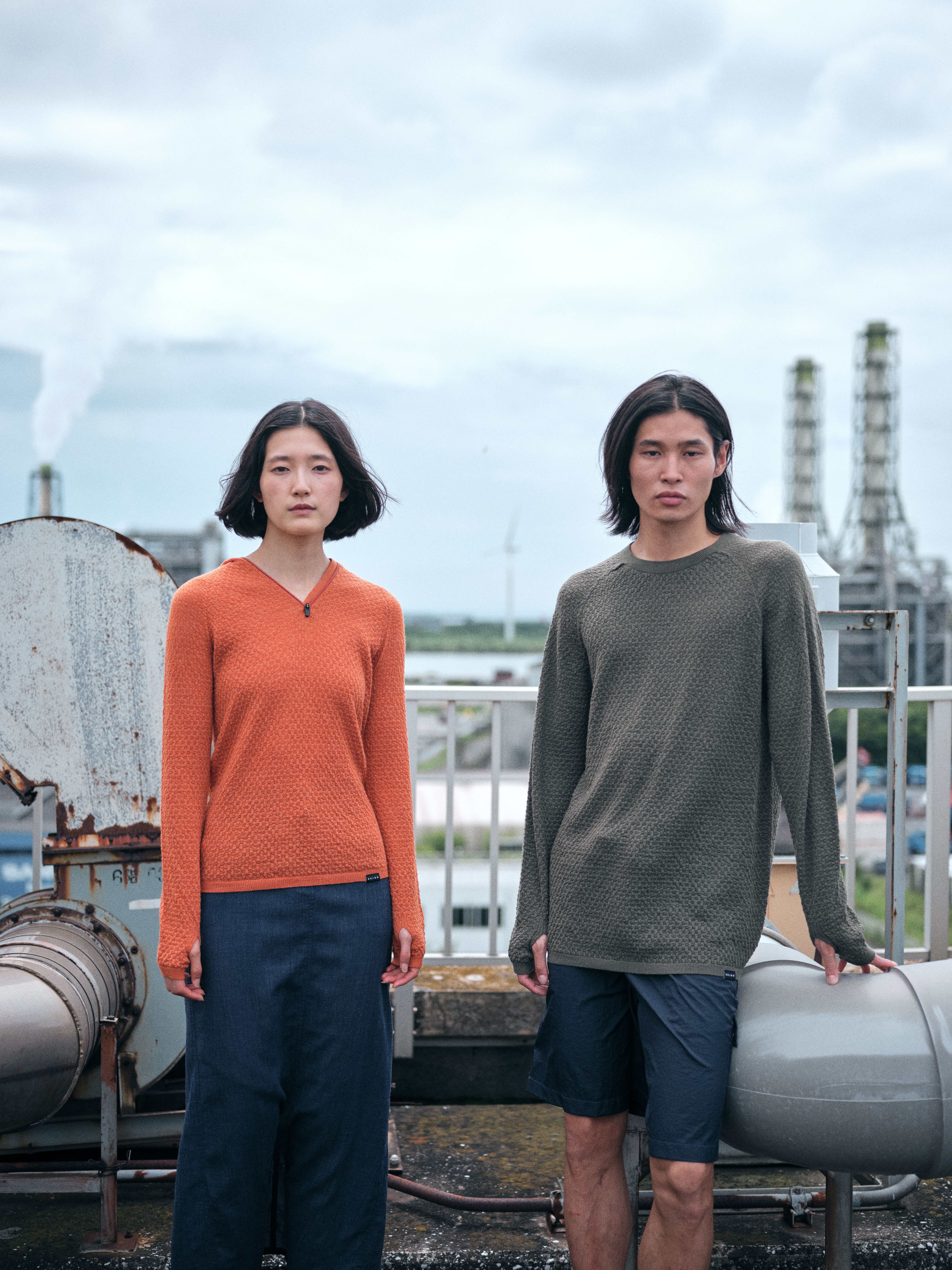 Two individuals standing on a rooftop, wearing sweaters against an industrial backdrop.