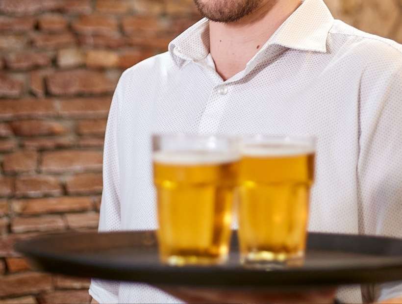 A waiter wearing a casual button down shirt and carrying two glasses of beer