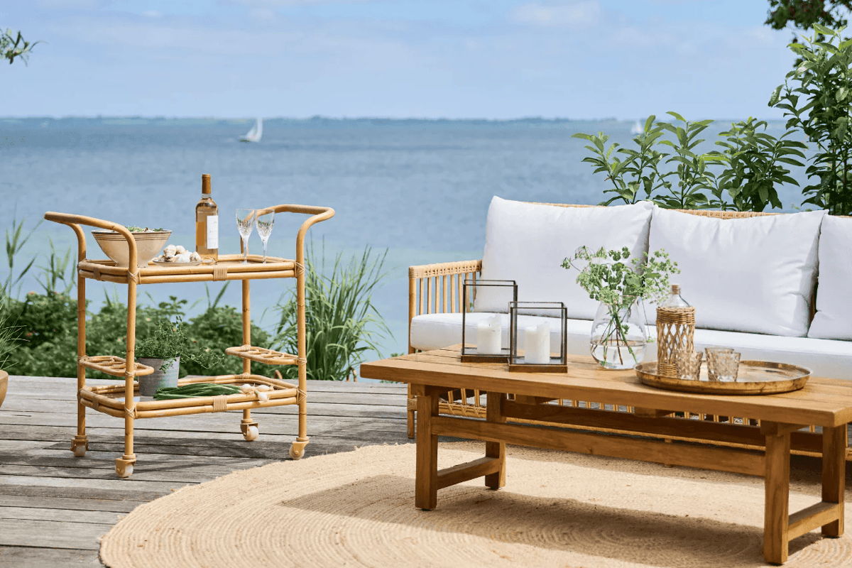 Coastal deck lounge with a white-cushion sofa, wooden coffee table, jute rug, and a rattan bar cart with drinks overlooking the ocean.