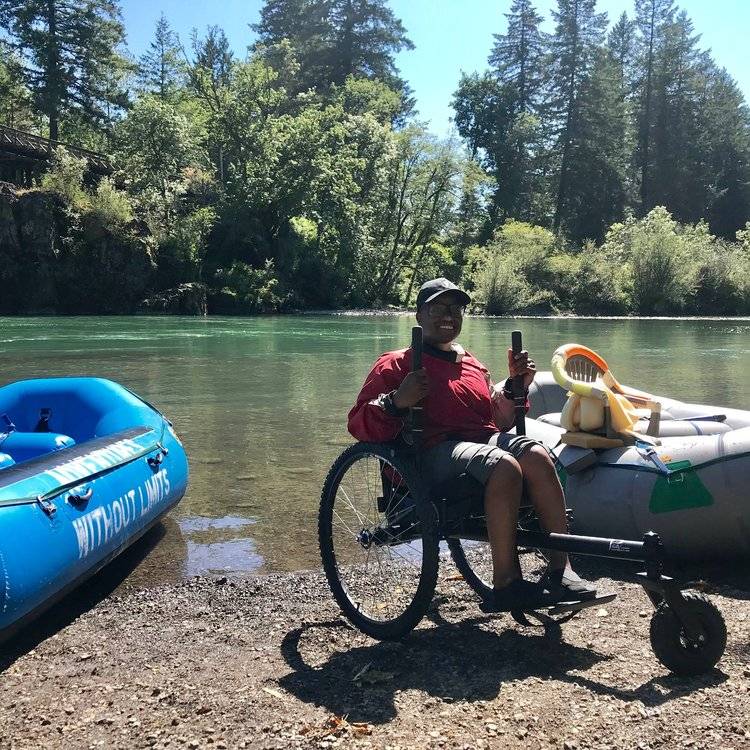 Person smiles using GRIT Freedom Chair off road wheelchair on gravel and dirt by kayaks partially landed and in water
