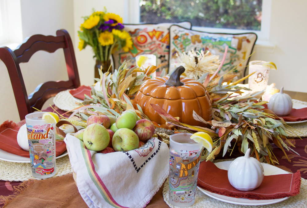 A fall table with pumpkins, apples, and fall table linens along with catstudio Geography glasses, dish towels, and pillows