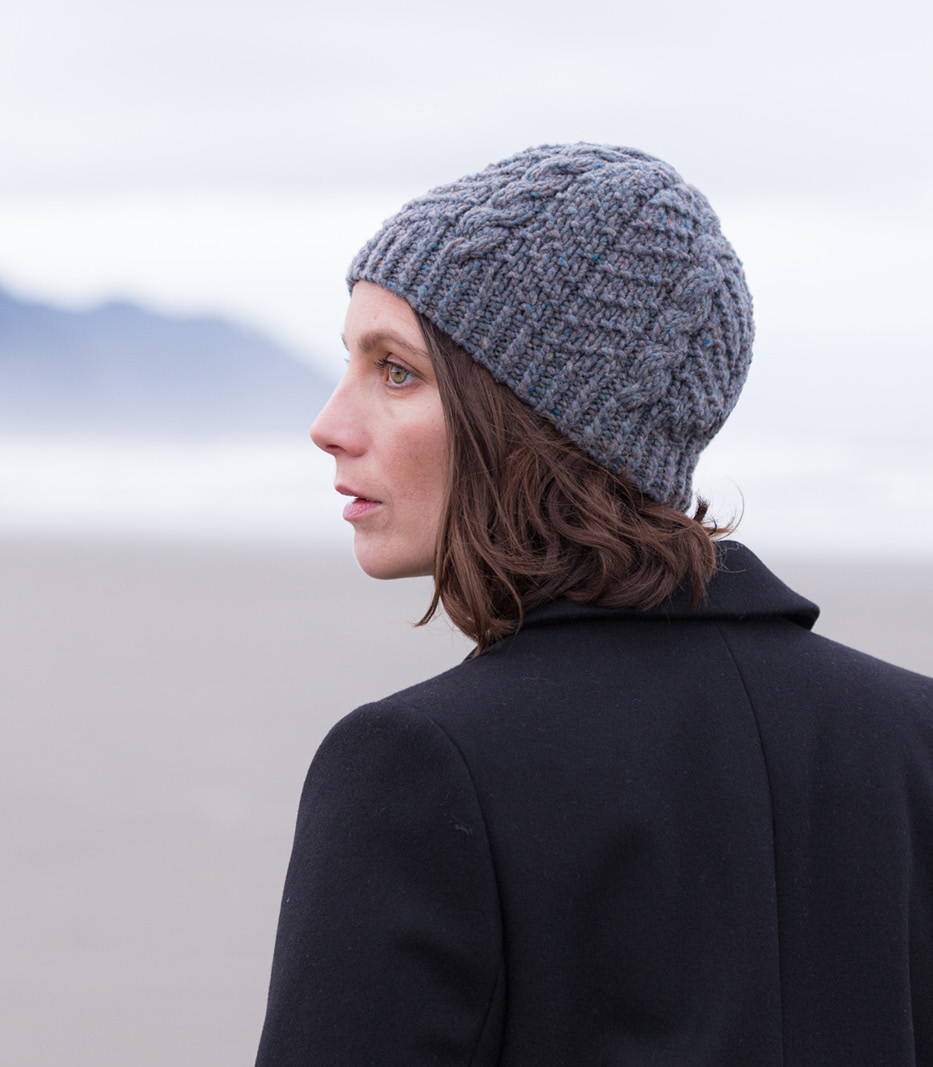 A woman on the oregon coast looking off to the left models a hand knit gansey cap in a chunky heathered yarn.