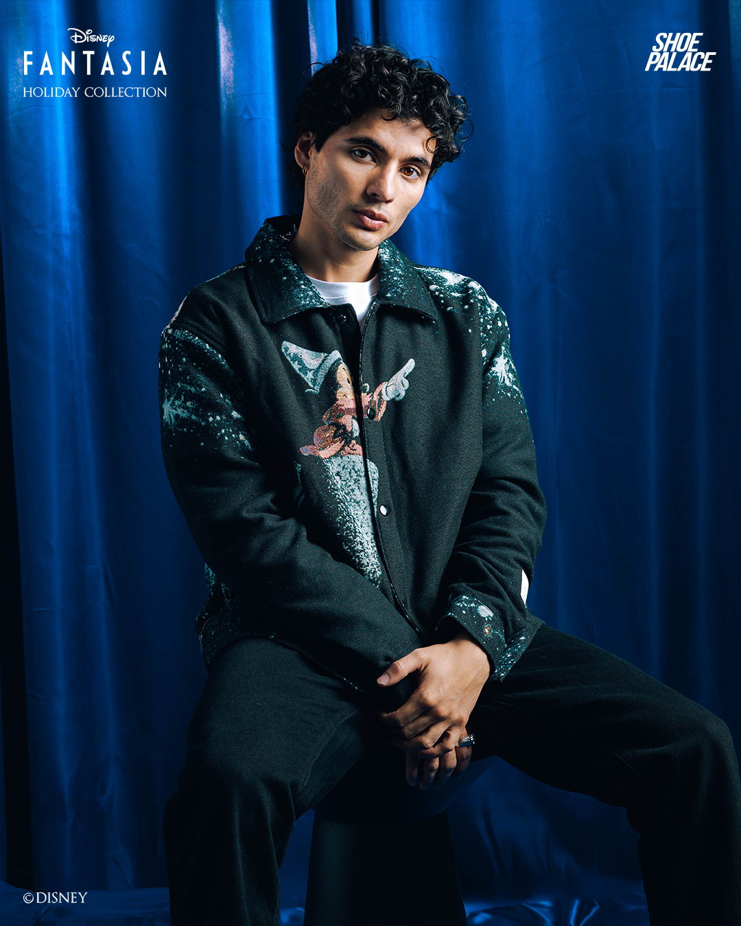 Young man sitting in front of a blue curtain, wearing a stylish jacket.