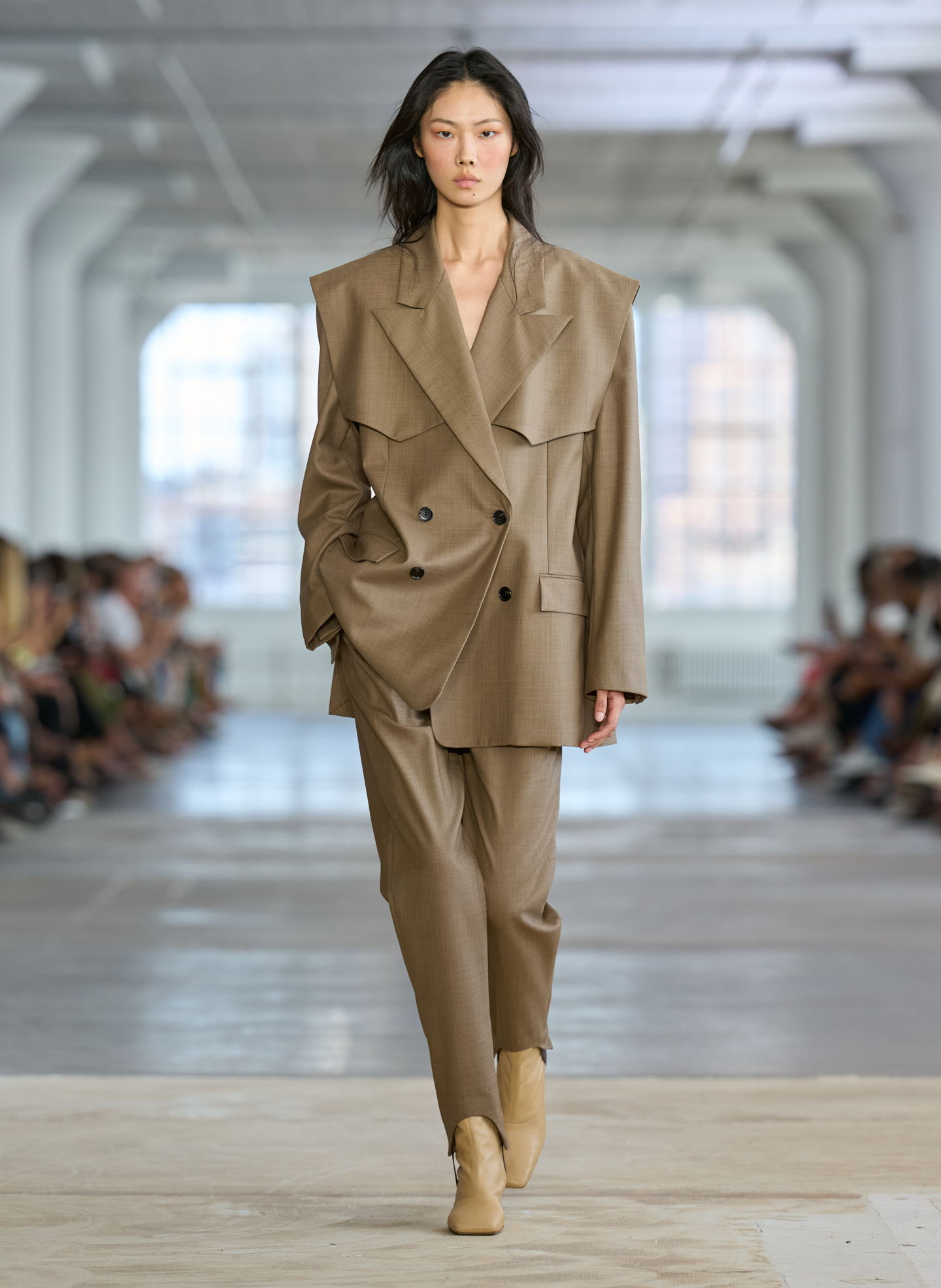 Model walking on runway in oversized brown suit and beige shoes.