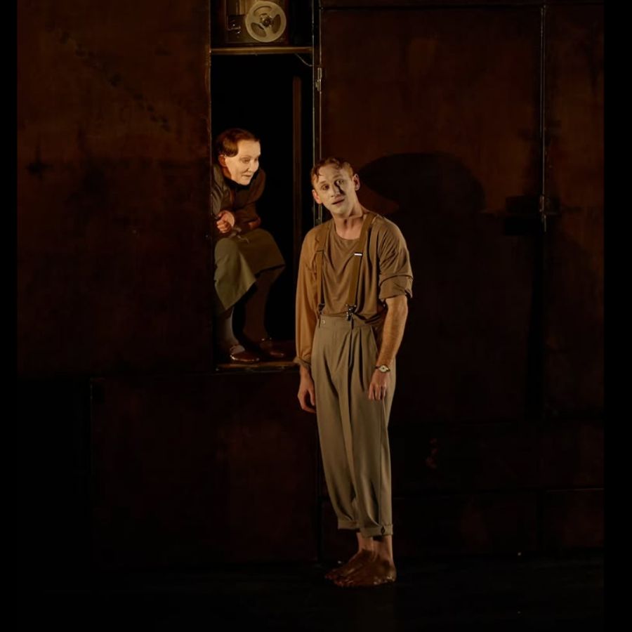Two performers on stage, one peering from a doorway, dramatic lighting.