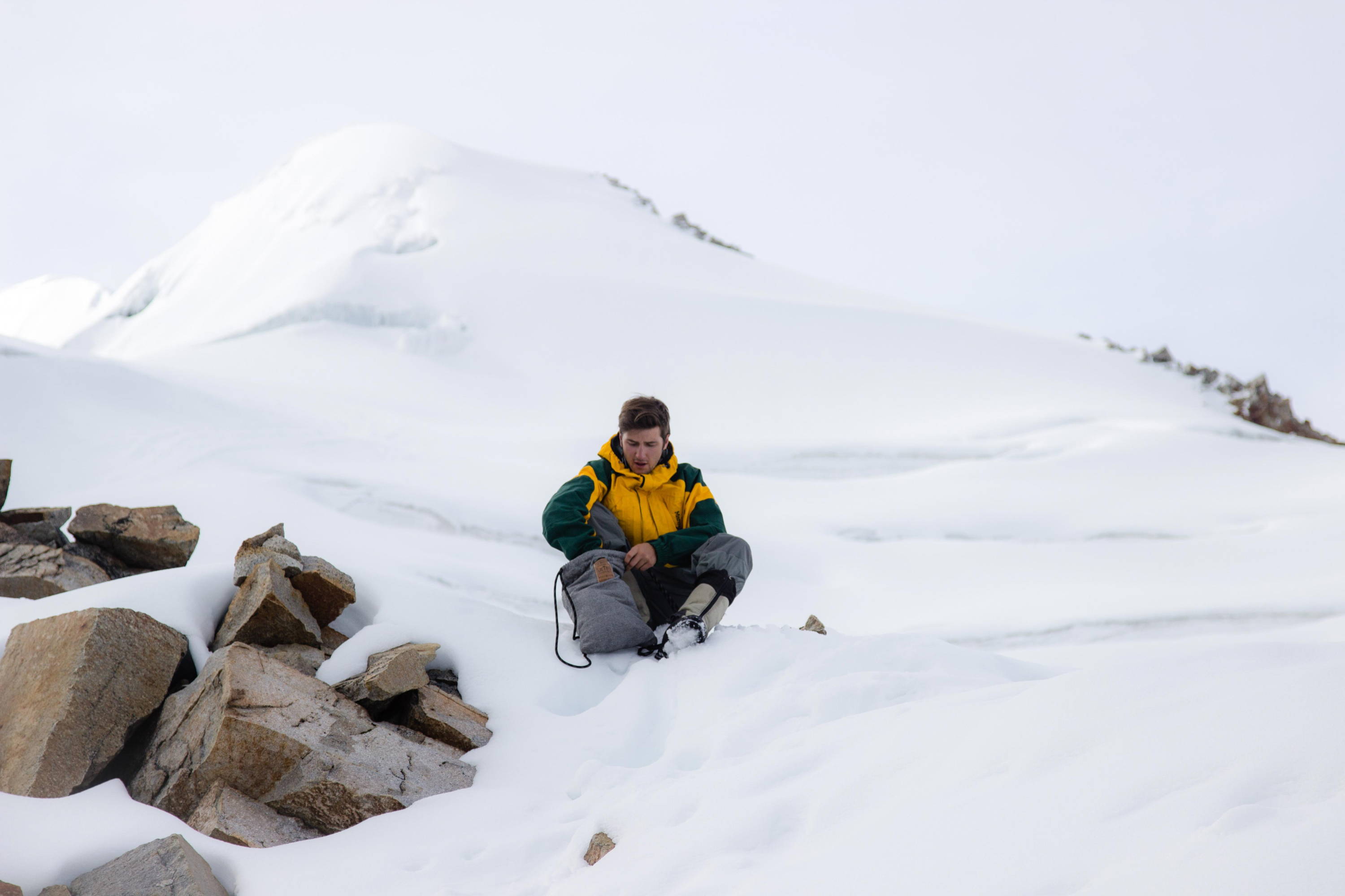 male snowboarder wearing yellow jacket unpacking his anti-theft lockable bag sitting in snow