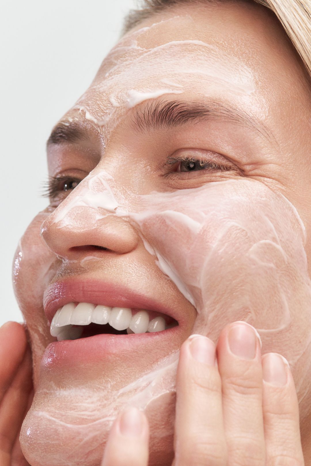 Woman applying facial cleanser, smiling with bubbles on her face.