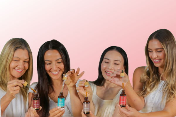 Four women holding bottles, smiling, against a pink background.