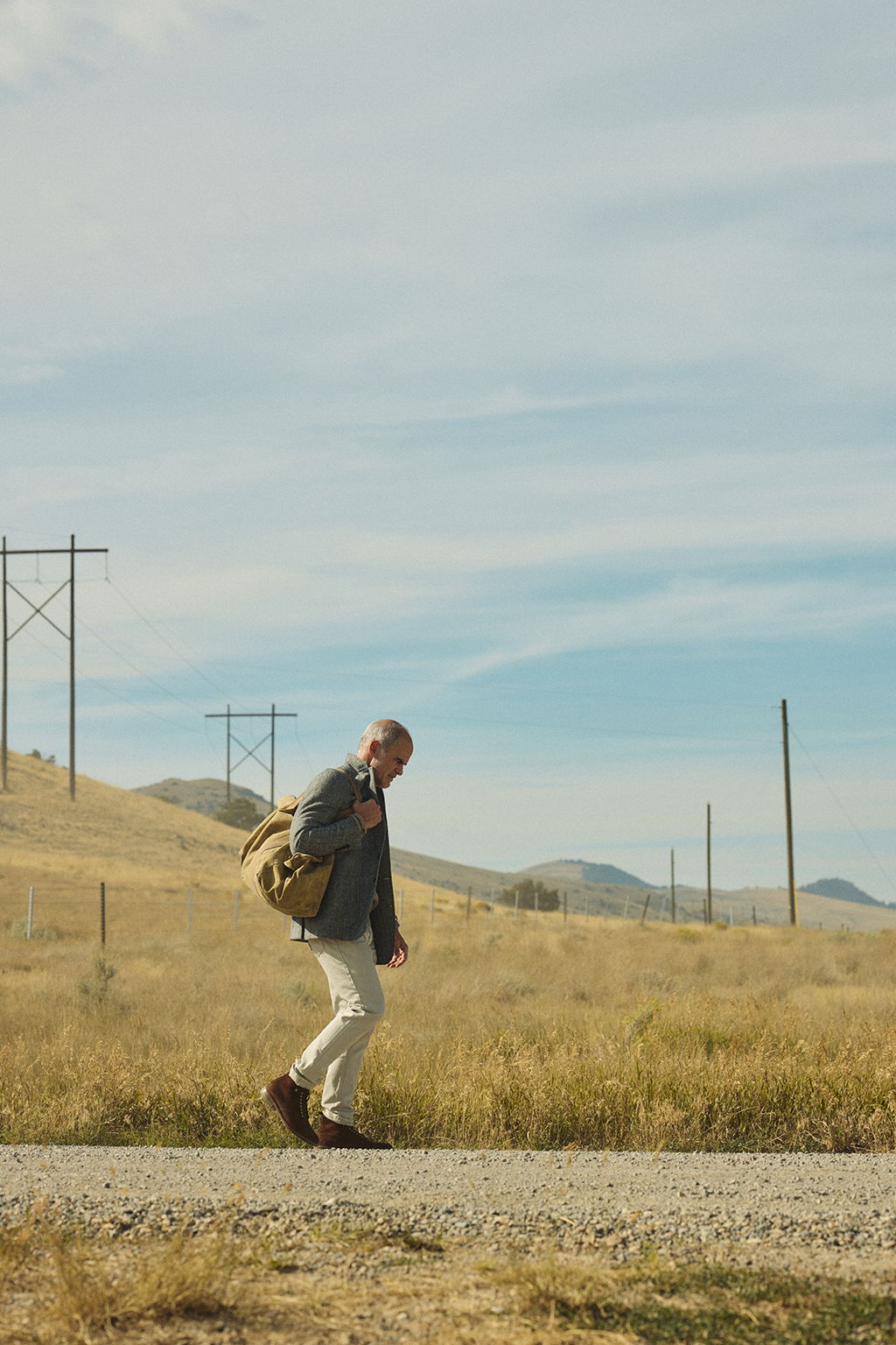Michael Kelly walking down a gravel road in wearing the Rivay Stanton Wool Herringbone Tweed Sportcoat over the Sonny Waffle Sweater in Khaki Marl with the WWade Garment Dyed Five Pocket Pant in Stone Grey holding a vintage air force pilots kit bag in Emigrant, Montana