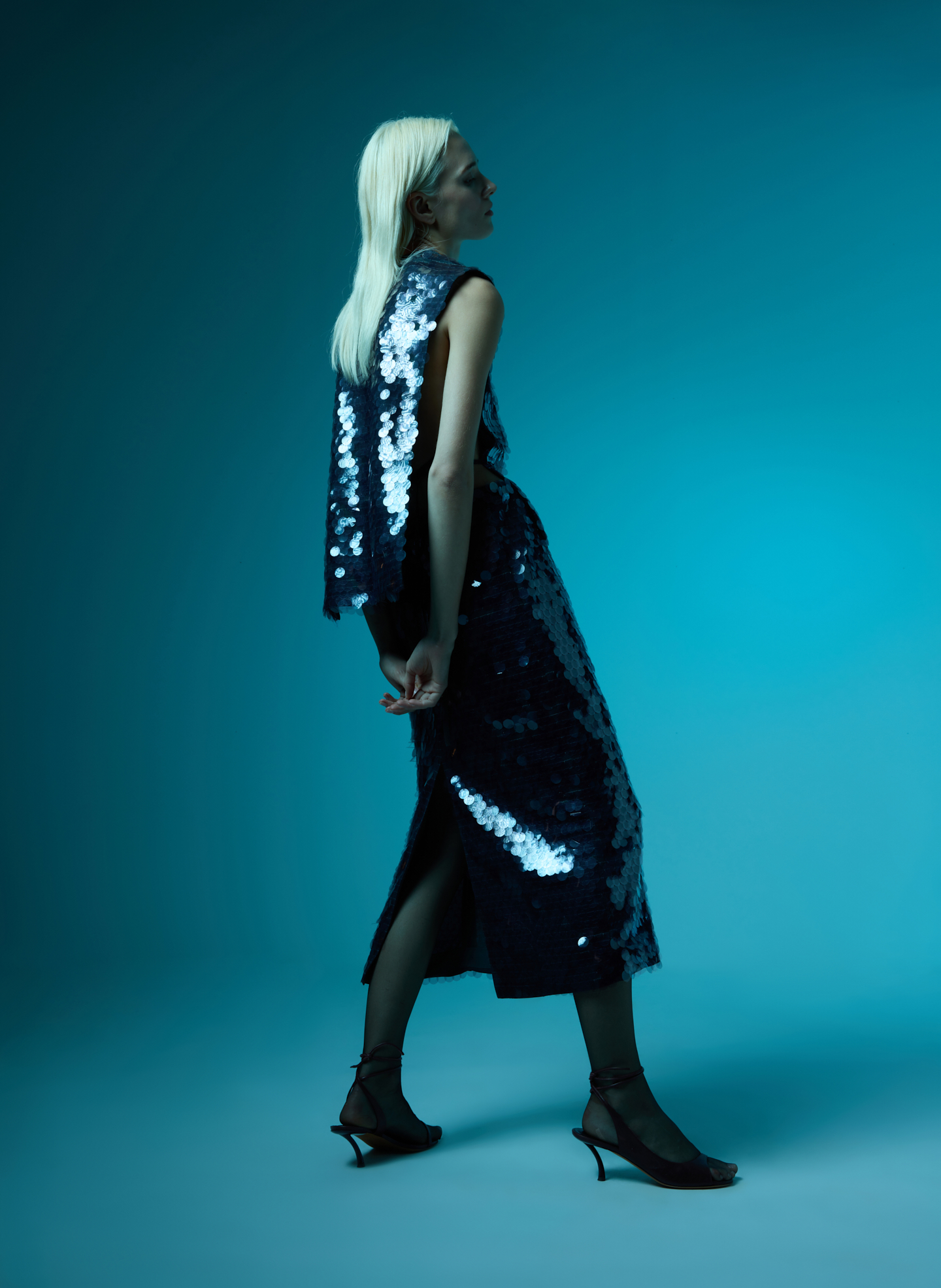 Model in sequined dress against a blue background, walking gracefully.
