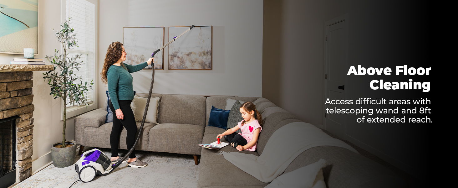 Woman cleaning above floor with vacuum; child sitting nearby on couch.