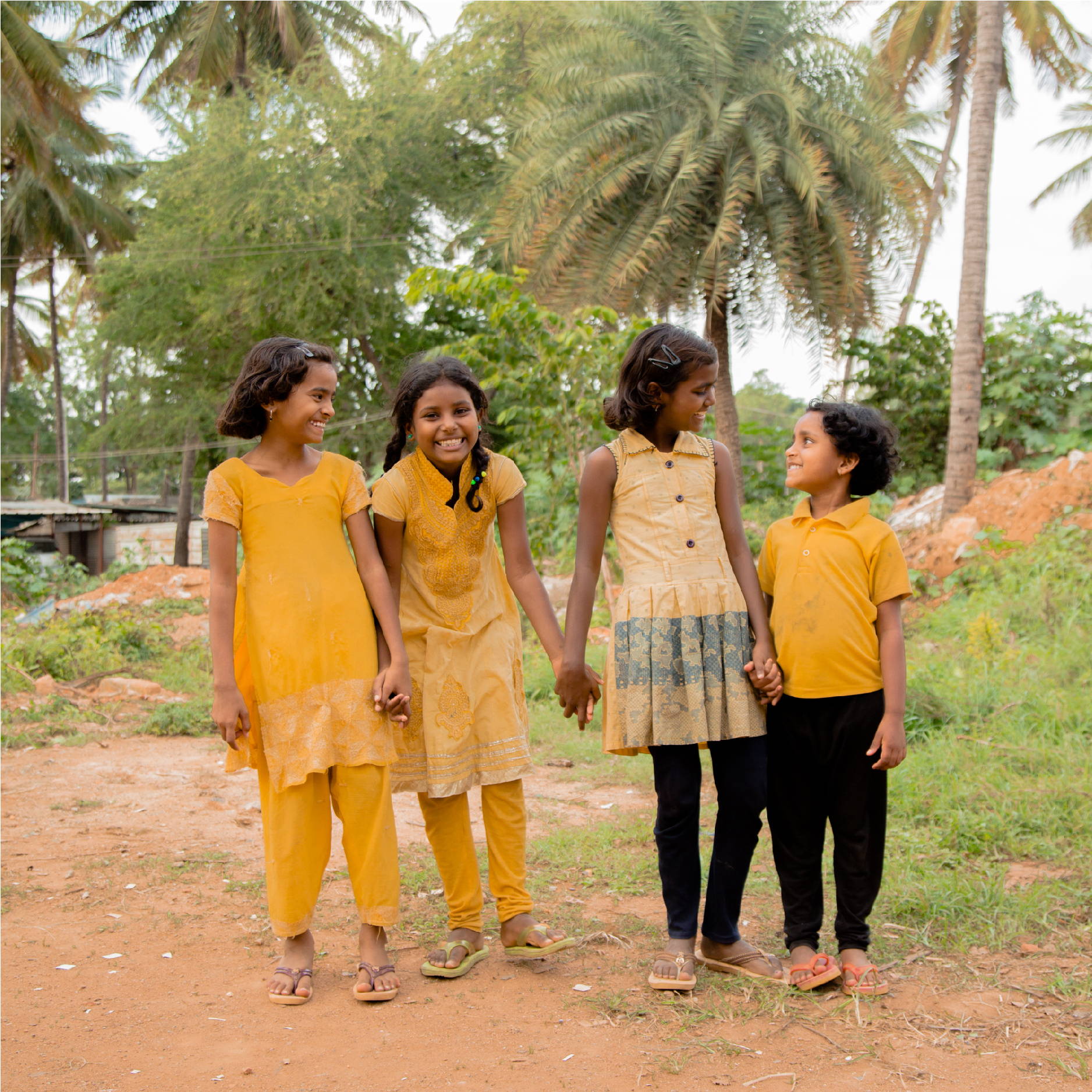 3 young Indian girls, hold hands while smiling, laughing, and looking at each other. They are all wearing yellow dresses.