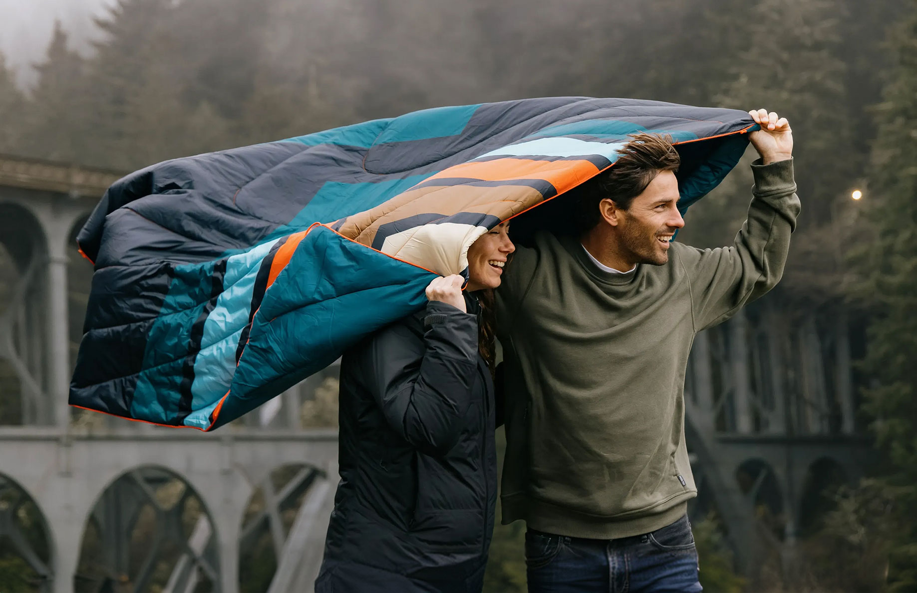 Two people under a blanket with a scenic background.