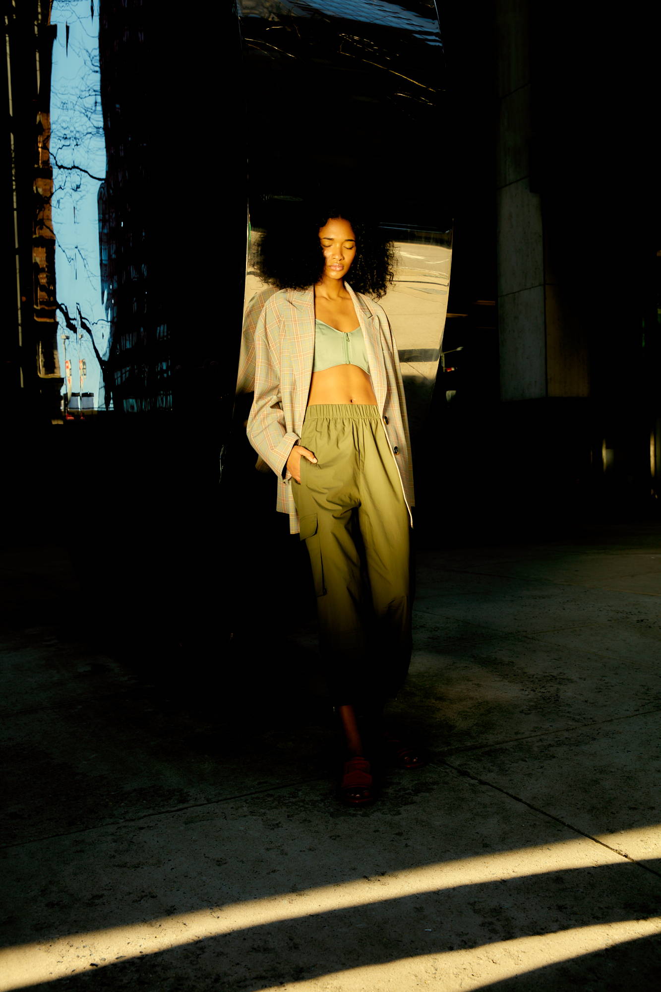 A person in stylish clothing stands in a sunlit urban setting.