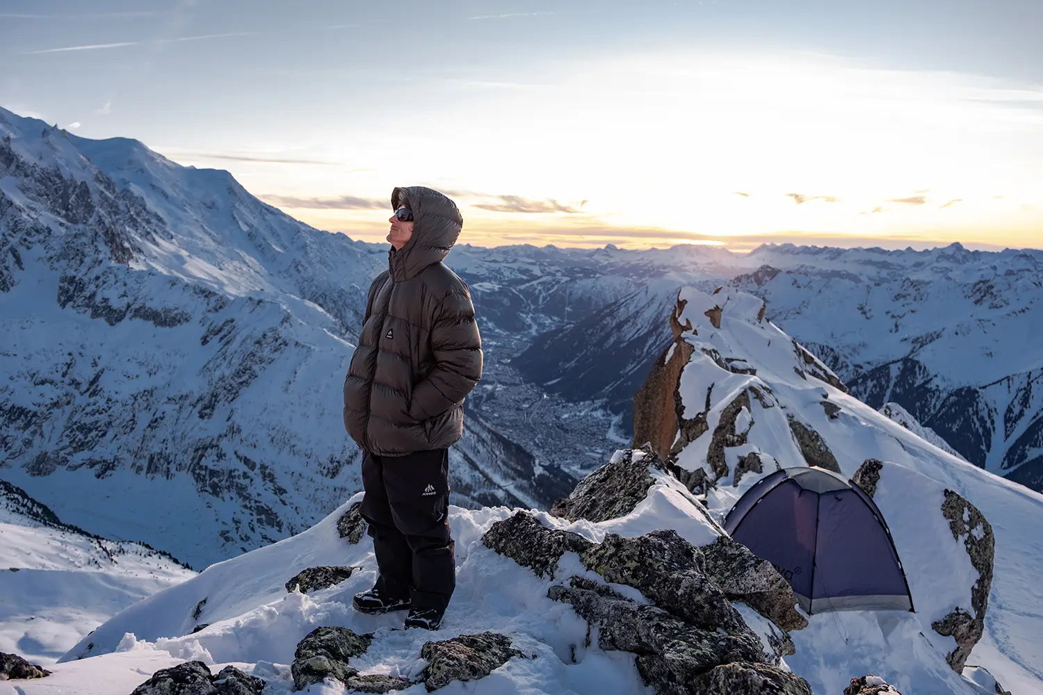 Person standing on a snowy mountain with a tent at sunset.