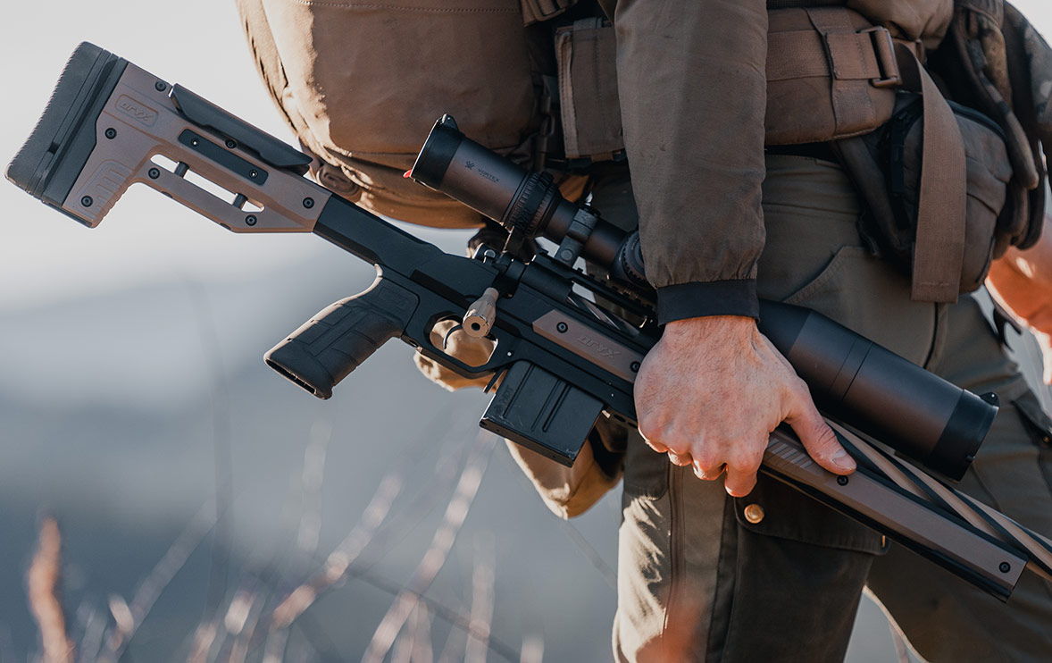 Person holding a rifle with a scope in an outdoor setting.