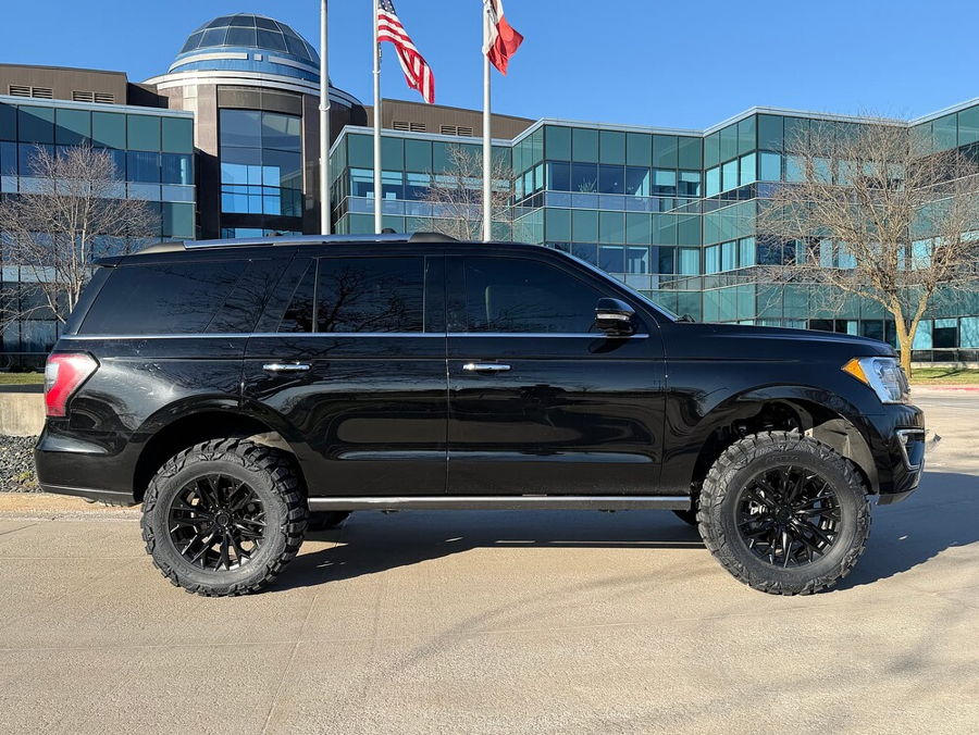 Side profile Troy's black 2018 Ford Expedition with off-road wheels parked before modern glass office building with flags gallery photo 5