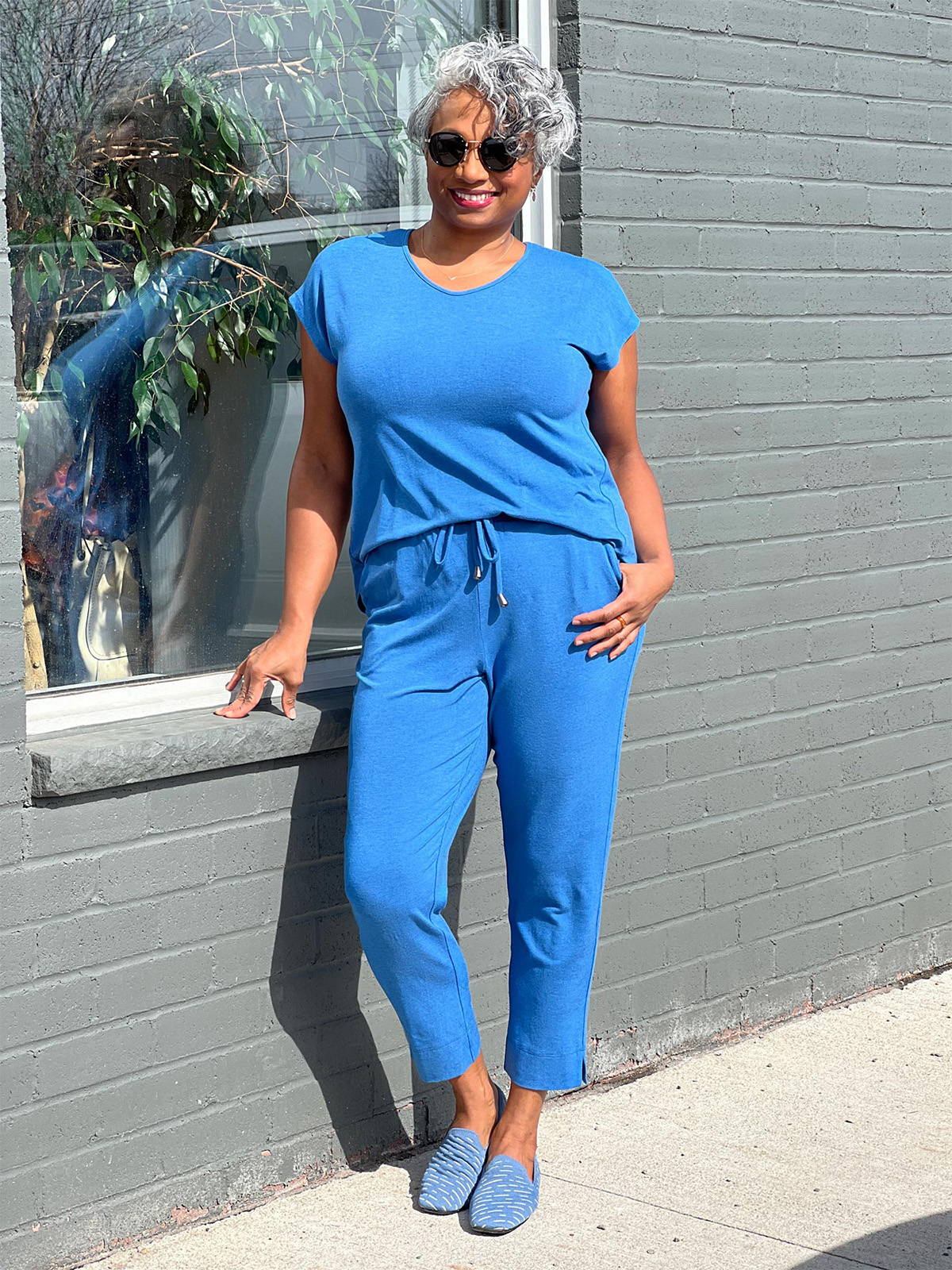 Woman standing next to a window smiling wearing Miik's Stef v-neck open-back capri jumpsuit in blue.