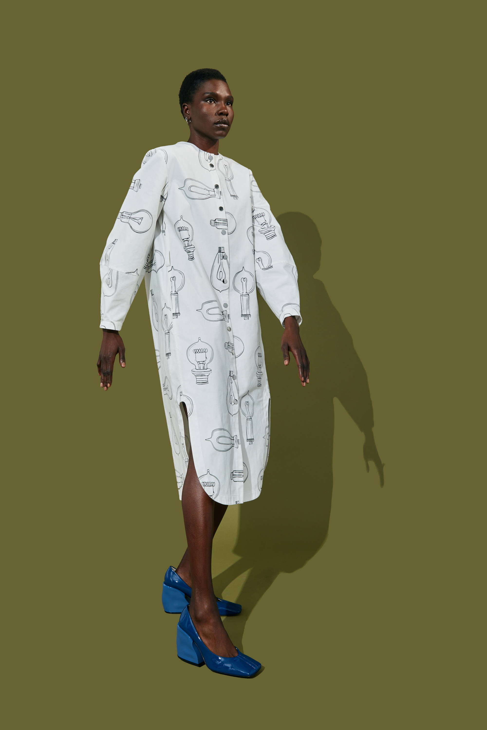 Model wearing a white dress with patterns and blue shoes against a green background.