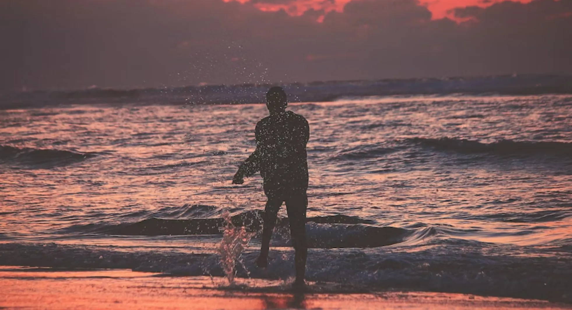 A person splashes along the shore against a red sky with clouds.